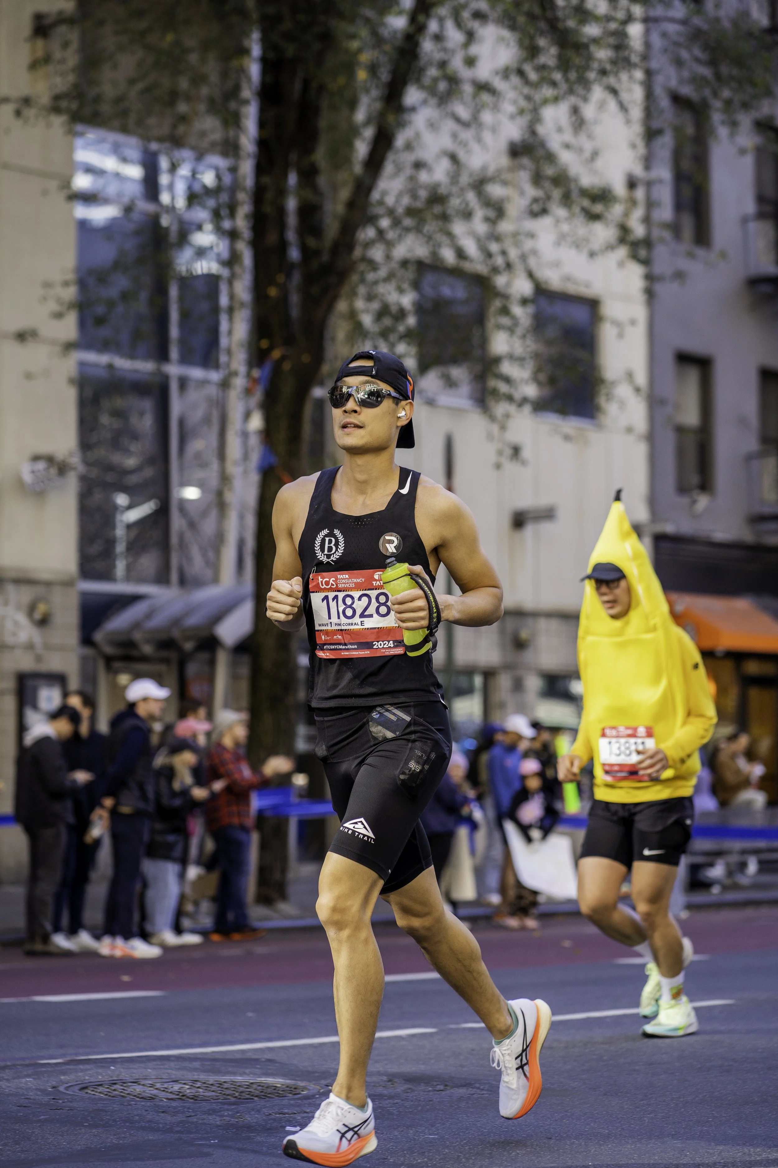 A male marathon runner in black athletic gear, sunglasses, and a backward cap, running on a city street during a race, with spectators and another runner in a yellow costume in the background.