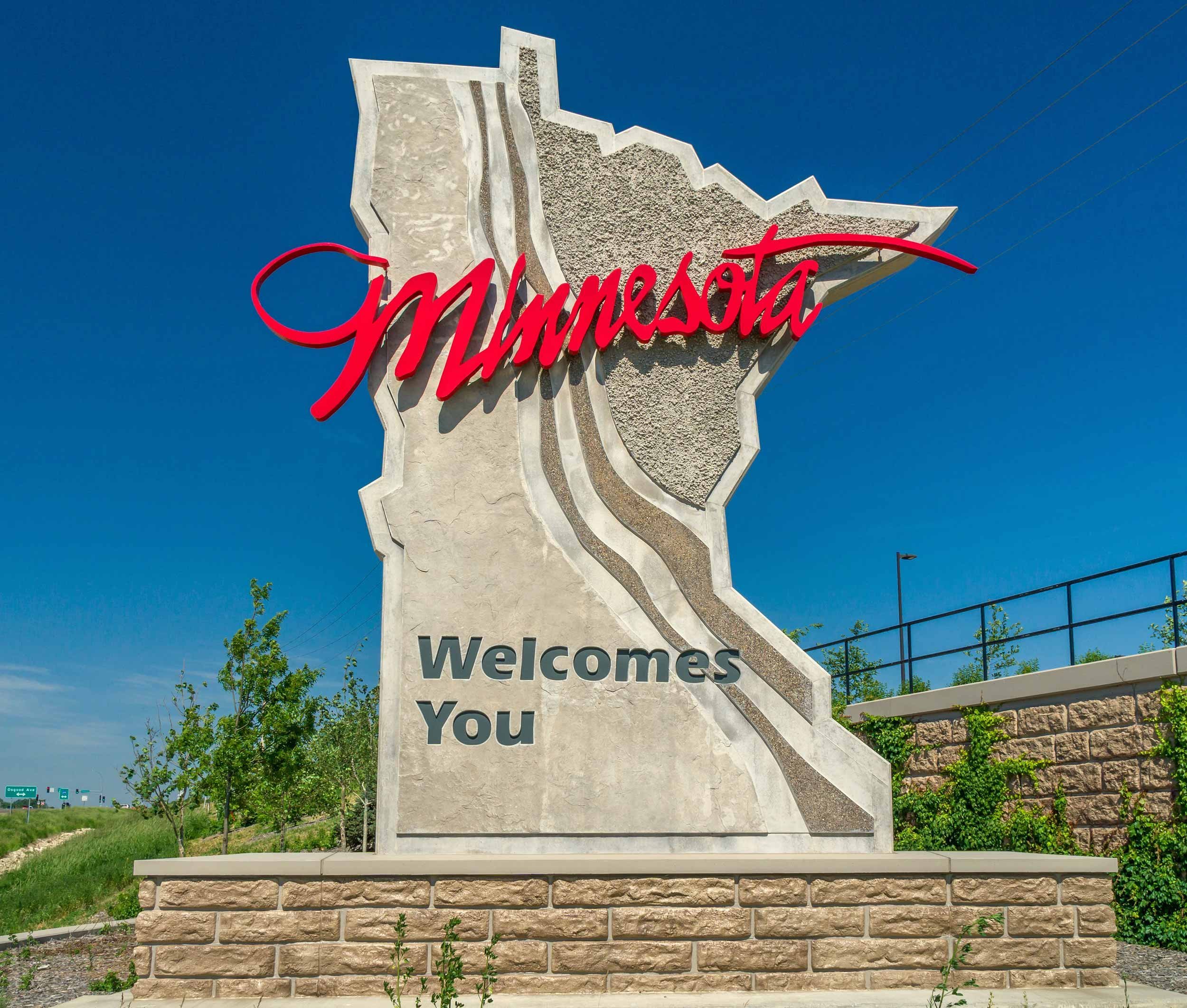 A welcome sign featuring the state of Minnesota with the word 'Minnesota' in red cursive overlaying the shape, and 'Welcome' in black text at the bottom. The sign is set against a clear blue sky.