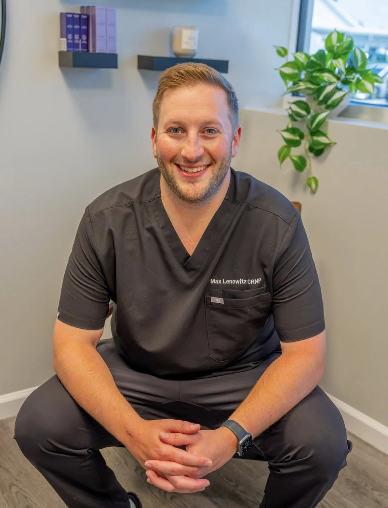 A smiling man wearing black scrubs with embroidered name Max Lenowitz, sitting on a chair in a room with gray walls, a window, a green leafy plant, and wall-mounted purple and gray folders.
