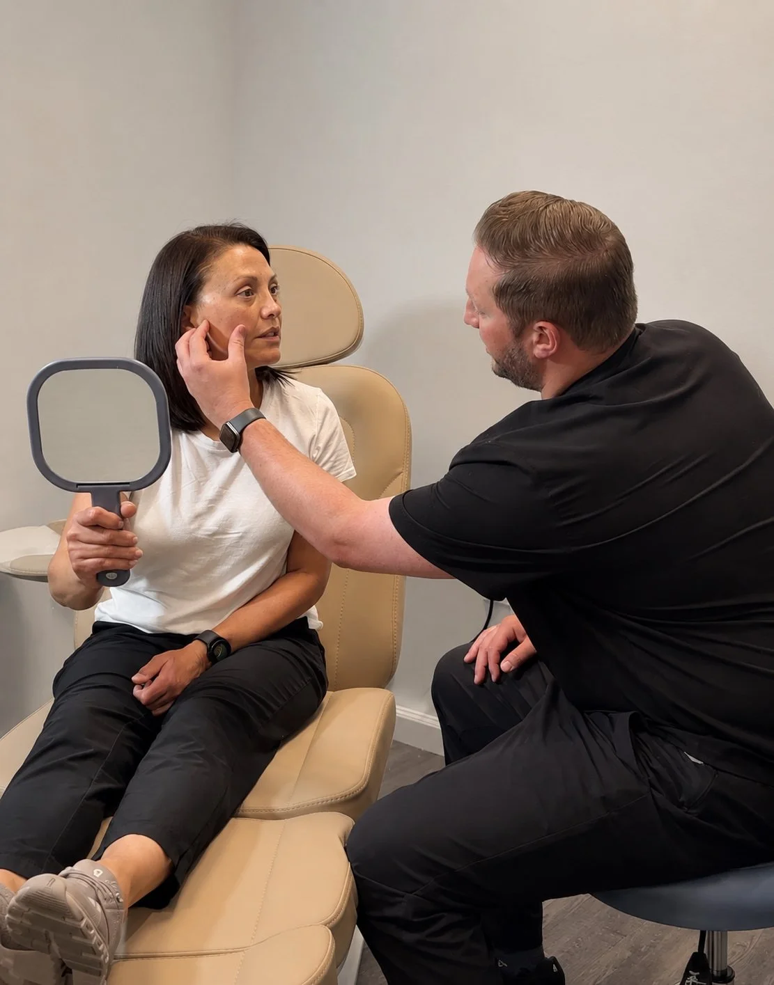 A woman sitting on a medical examination chair holding a mirror, while a man in black pokes her face to examine her skin.