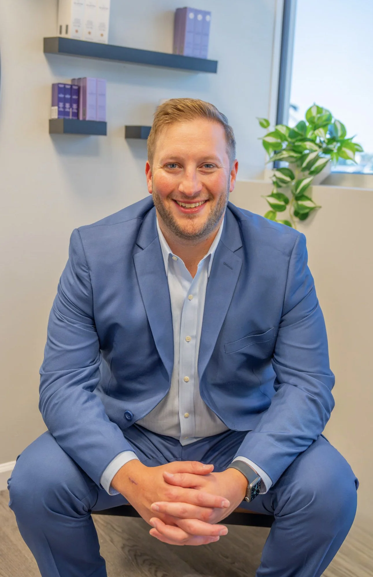 A young man in a blue suit sitting cross-legged on the floor, smiling at the camera in an office setting with a window and green plant in the background.