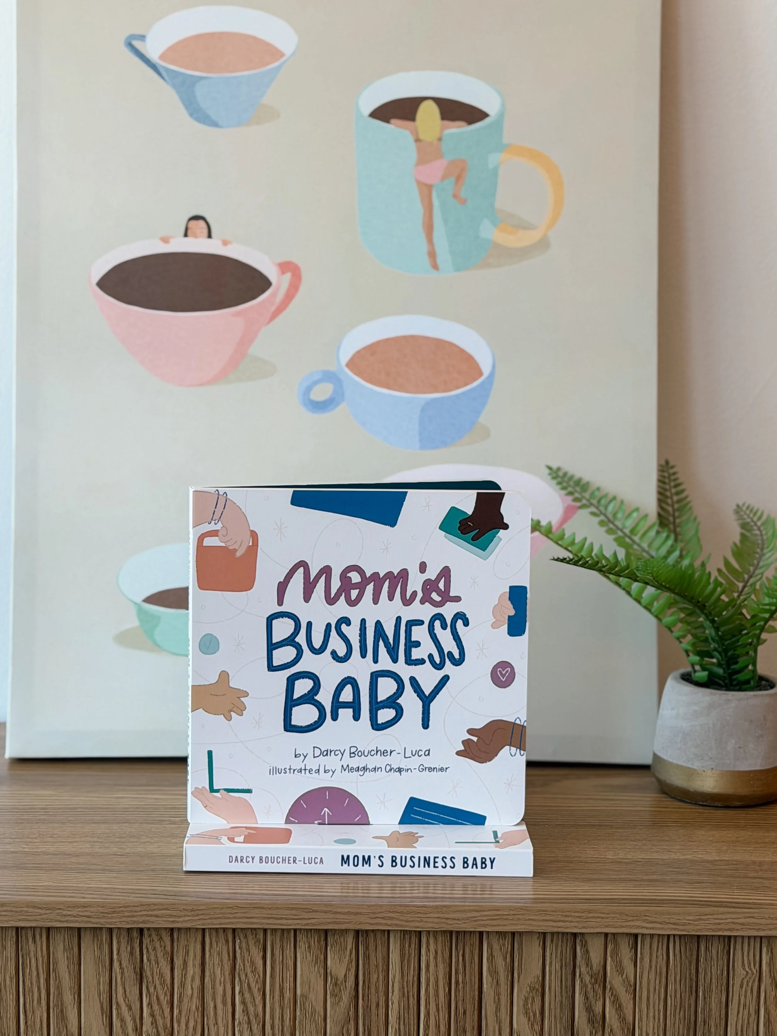 Book titled "Mom's Business Baby" by Darcy Boucher-Luca, illustrated by Meghand Chapin-Grenier, displayed on a wooden surface alongside a potted fern plant; background features a wall with illustrations of coffee cups and a mug with a figure of a woman in a pink swimsuit climbing out of coffee.