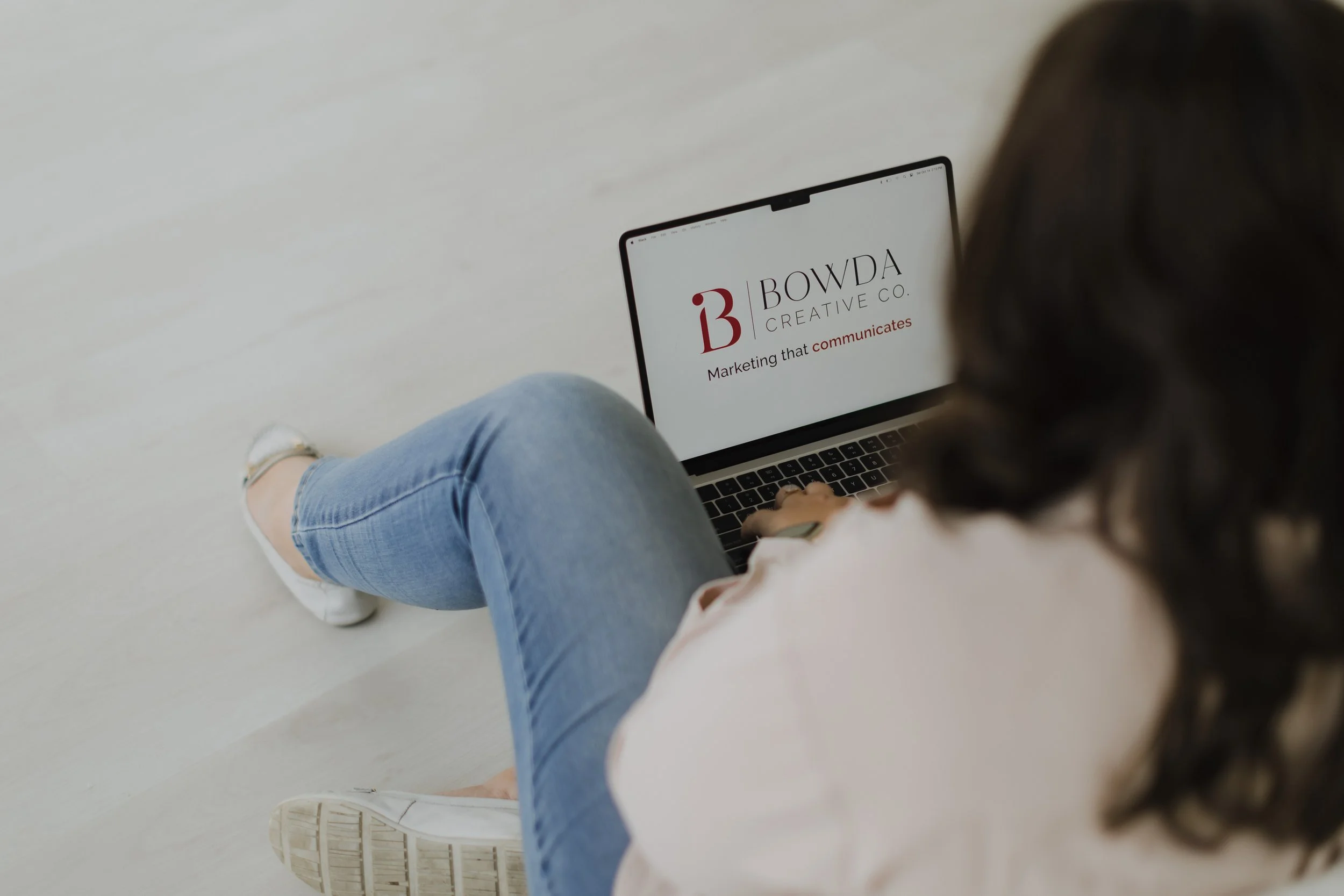 Person sitting on the floor using a laptop with the screen displaying 'BOWDA CREATIVE CO' and the tagline 'Marketing that communicates.'