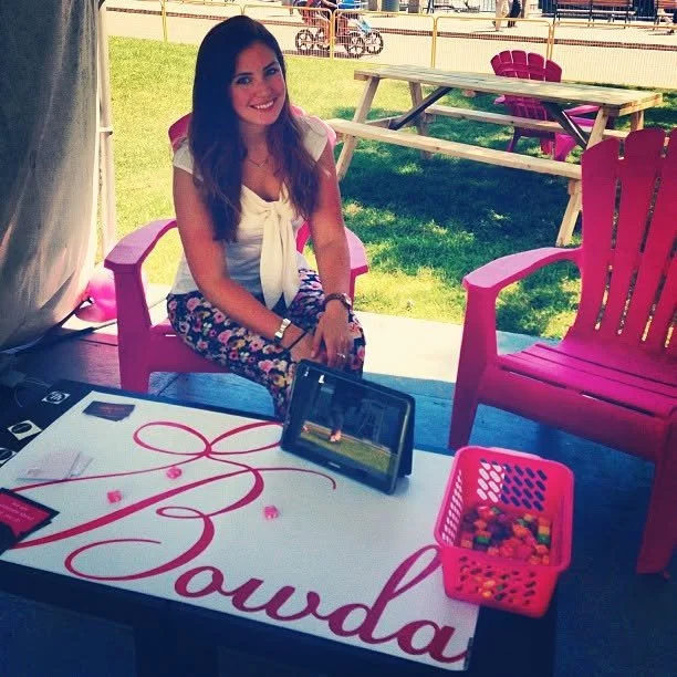 Young woman sitting under a tent at a table with a pink and white sign that says 'Lo Budda,' a tablet, and a pink basket of colorful candies, outdoors on a sunny day.