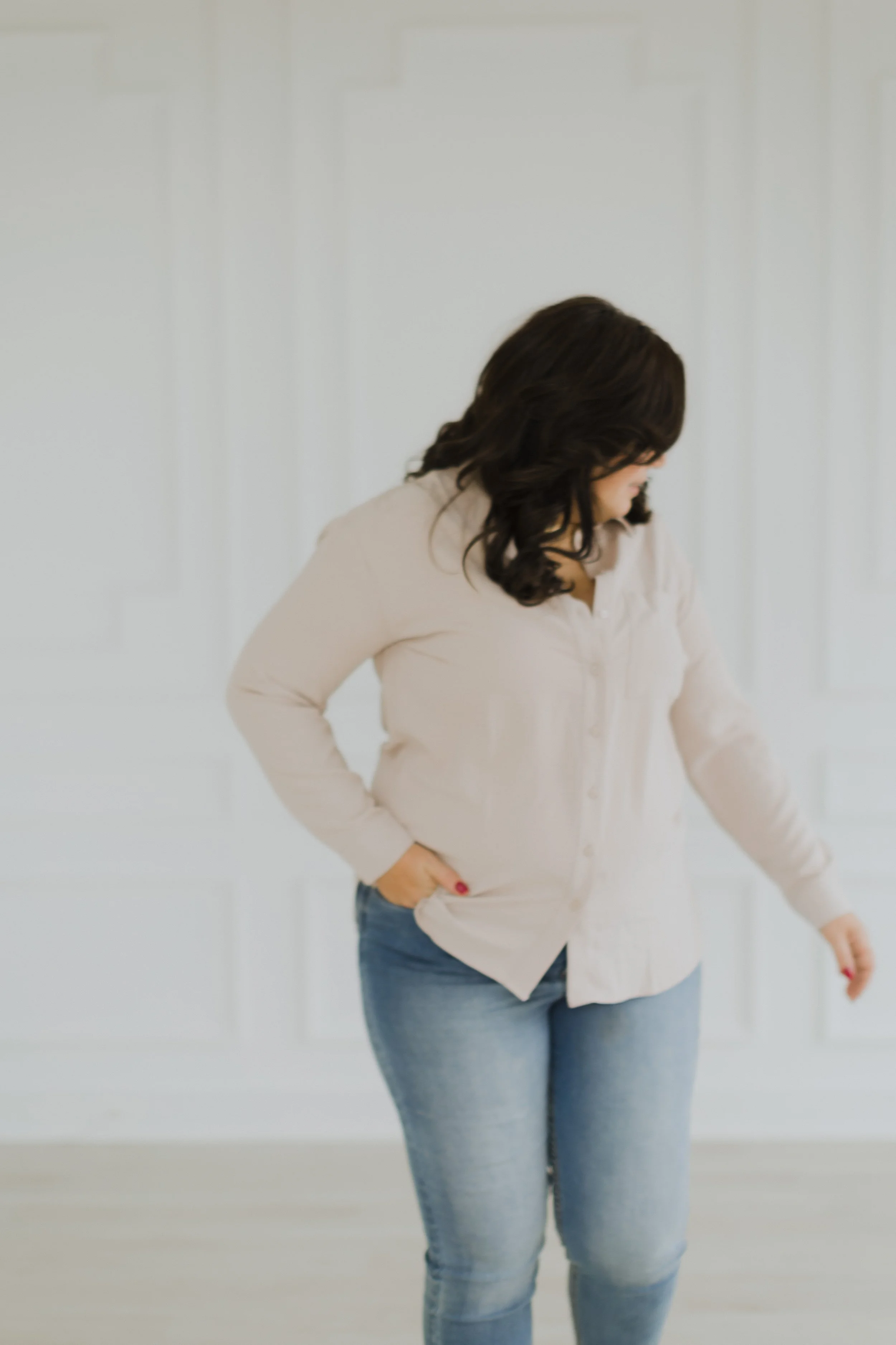 A woman with dark hair wearing a beige blouse and blue jeans, standing against a plain white wall.