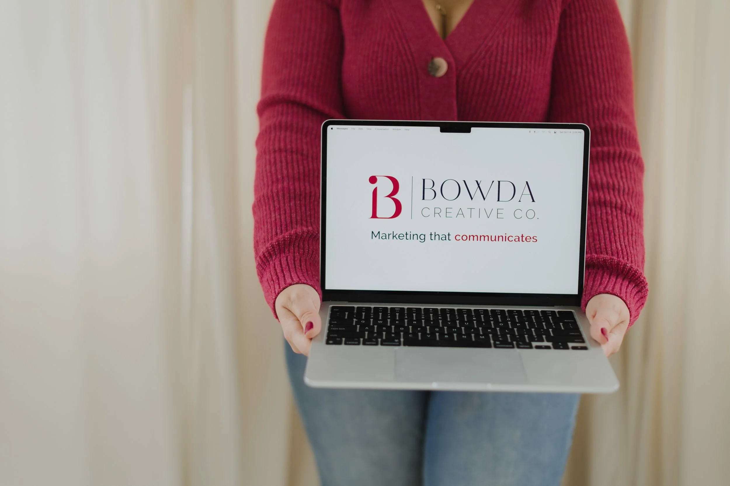 Person in a red sweater holding an open laptop displaying the logo and tagline of Bowda Creative Co.