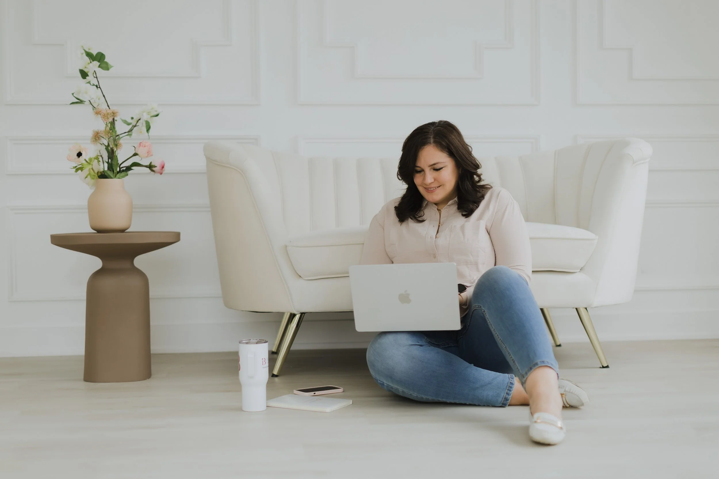 A woman with shoulder-length dark hair sitting on the floor in a bright, white room with decorative wall panels. She is smiling and working on a laptop, dressed in a light-colored blouse and jeans. Next to her is a beige side table with a tall beige vase holding a flower arrangement. In front of her on the floor are a white tumbler, a smartphone, and a closed notebook.