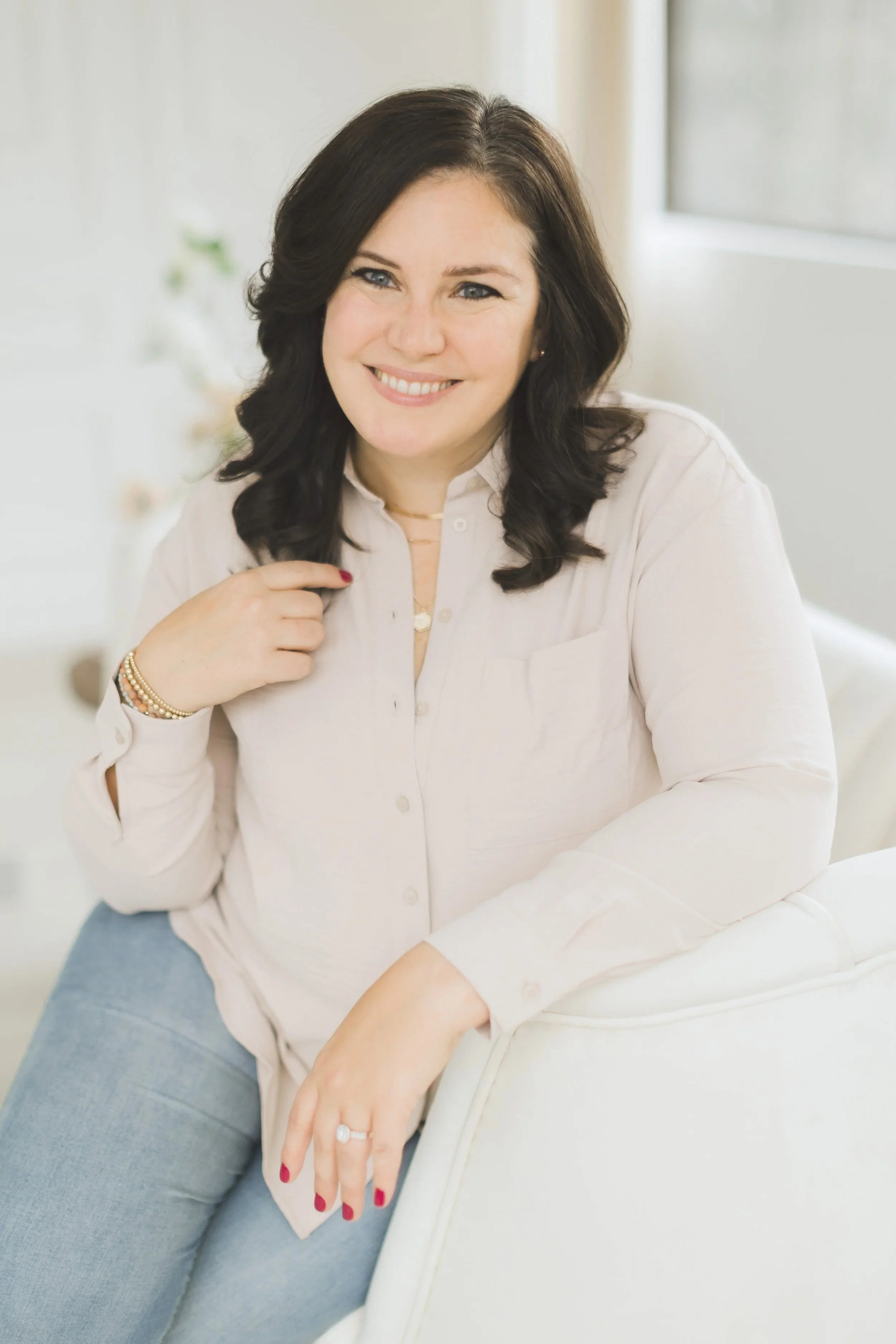 Portrait of a woman with dark hair, smiling, wearing a light pink shirt, light blue pants, sitting on a white chair in a bright room.