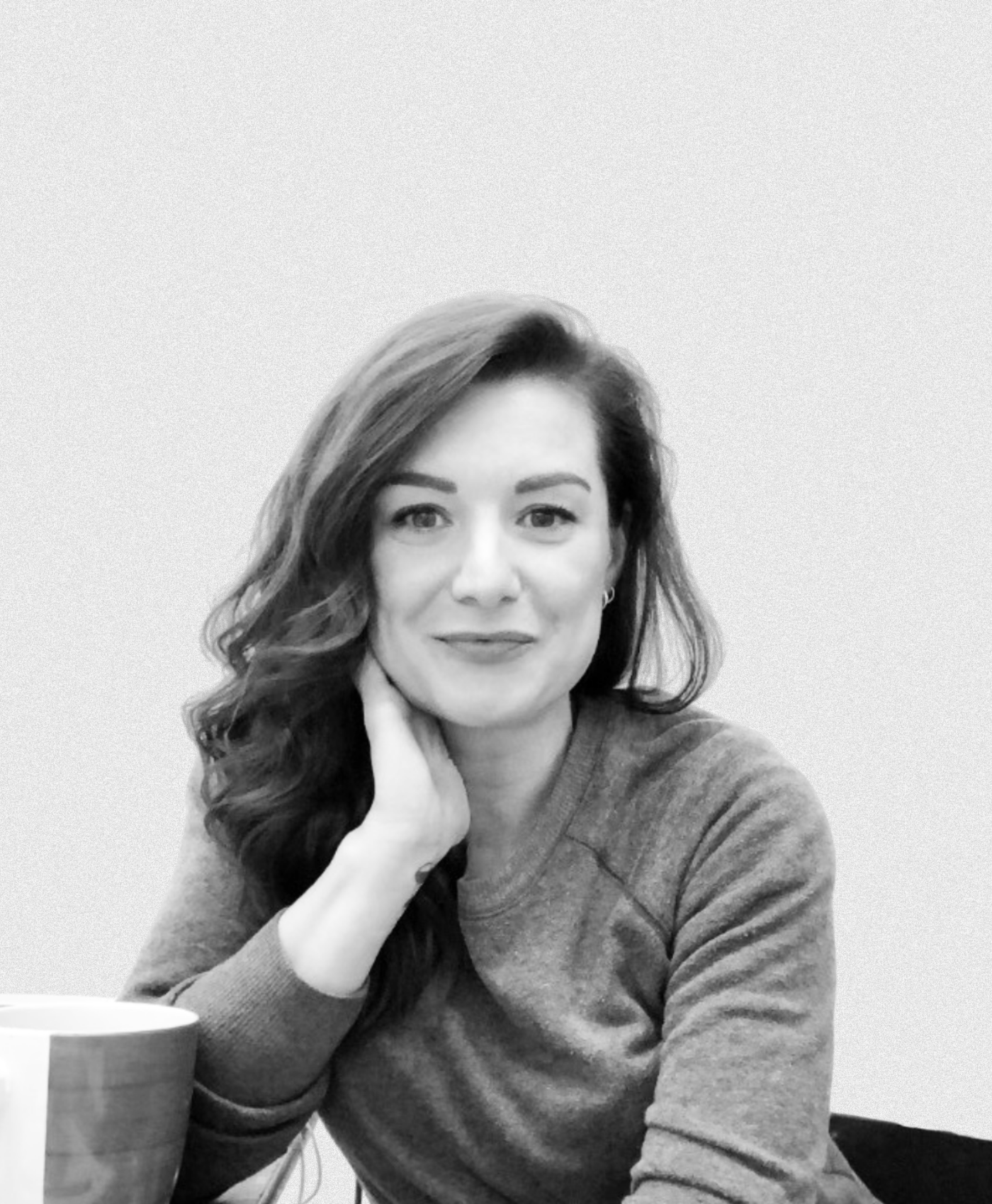 Black and white photo of a woman with long, wavy hair, sitting at a table with a mug in front of her. She is wearing a long sleeve shirt and is resting her chin on her hand, looking at the camera with a slight smile.