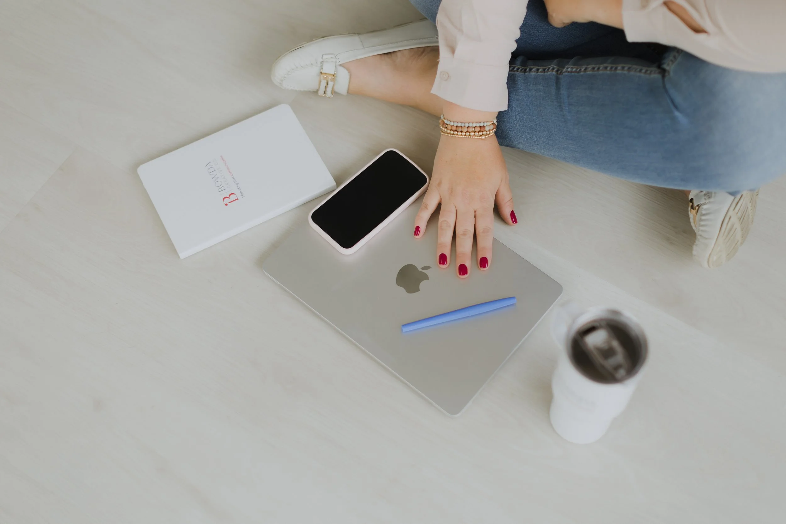 A person sitting on the floor with their legs crossed, wearing white sneakers, jeans, and bracelets. They have a white shirt and are using a silver laptop with a black smartphone placed on top of it. Nearby, there is a white book or notebook with red and black text, and a white cup with dark liquid.