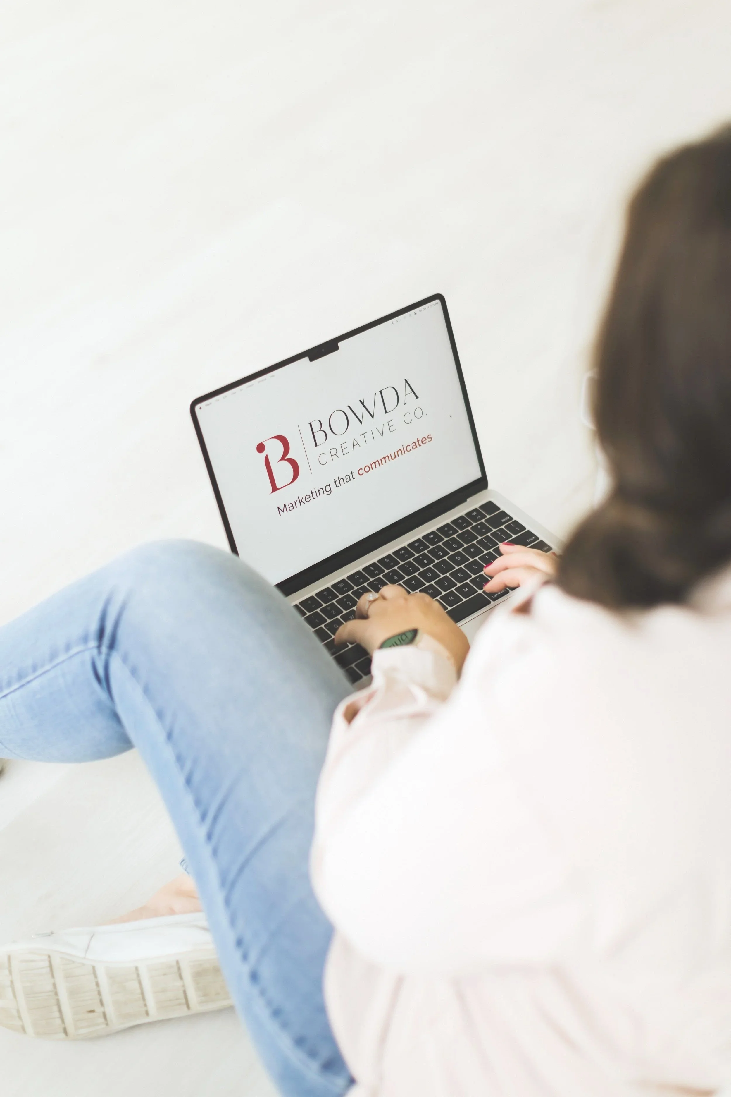 A person sitting cross-legged on a white surface, wearing light blue jeans and white sneakers, using a laptop with a screen displaying a logo and text.