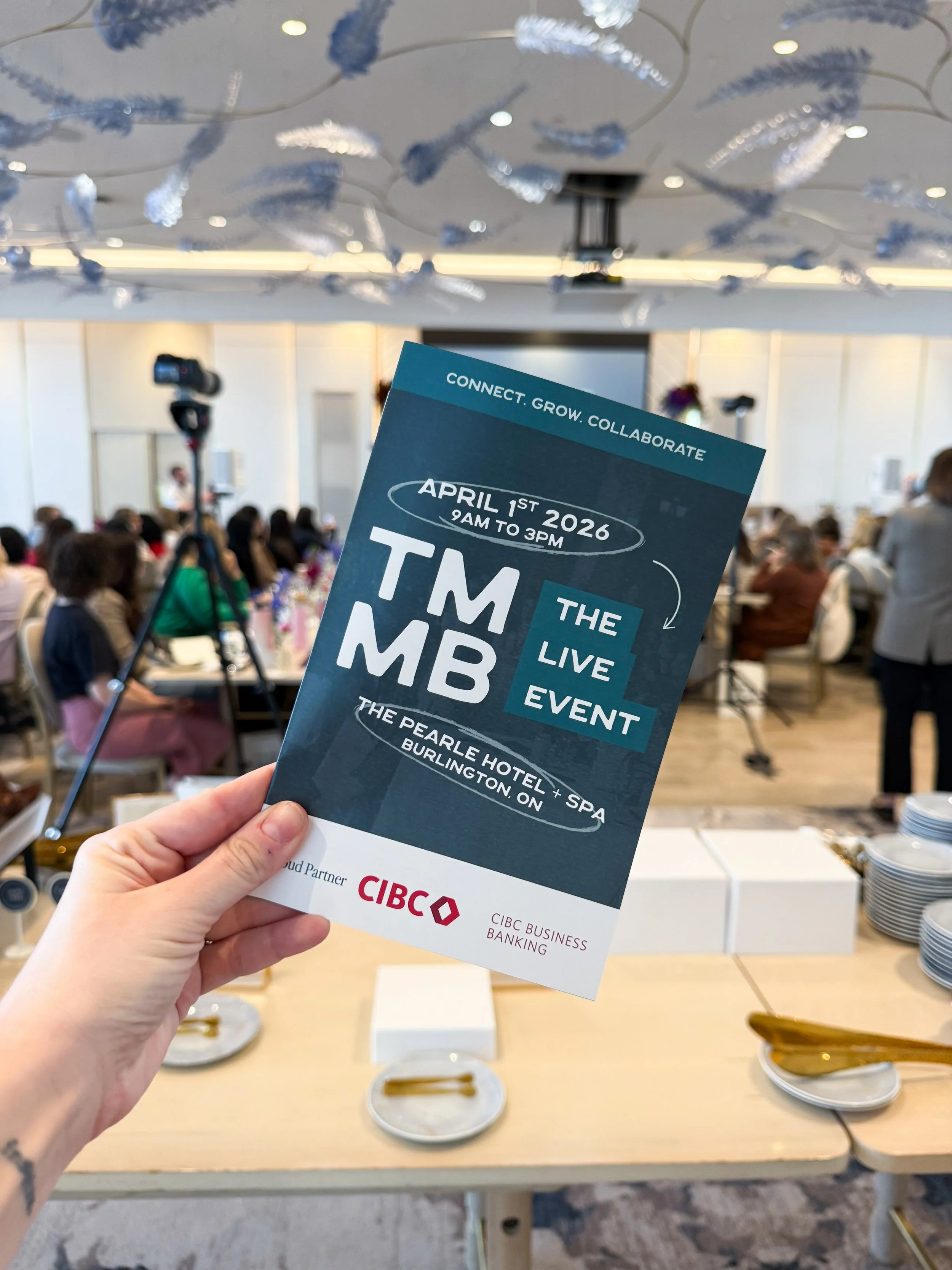 A person holding a flyer for 'The Live Event' of 'TMT' scheduled for April 1st, 2026, from 9 AM to 3 PM at The Pearle Hotel + Spa in Burlingtion, ON. The background shows a conference room with seated attendees, a camera on a tripod, and decorative ceiling ornaments.