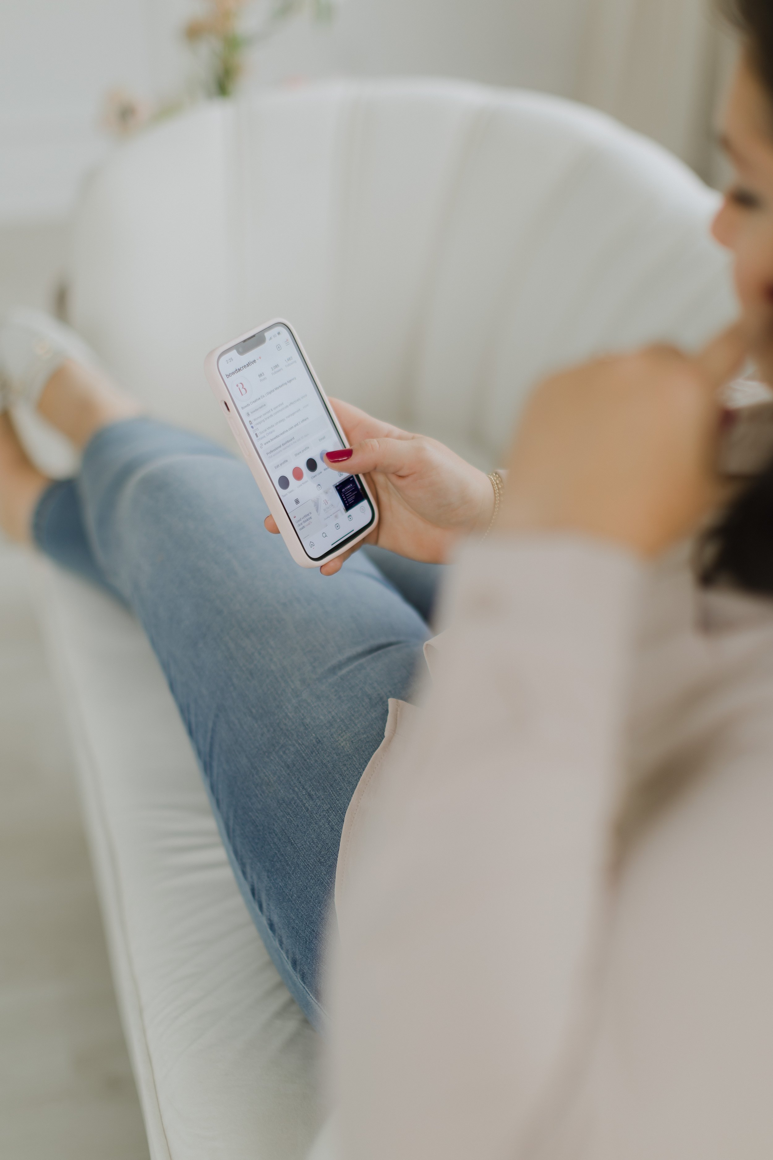 A woman sitting on a white chair, holding a smartphone in her right hand with a blurred background.