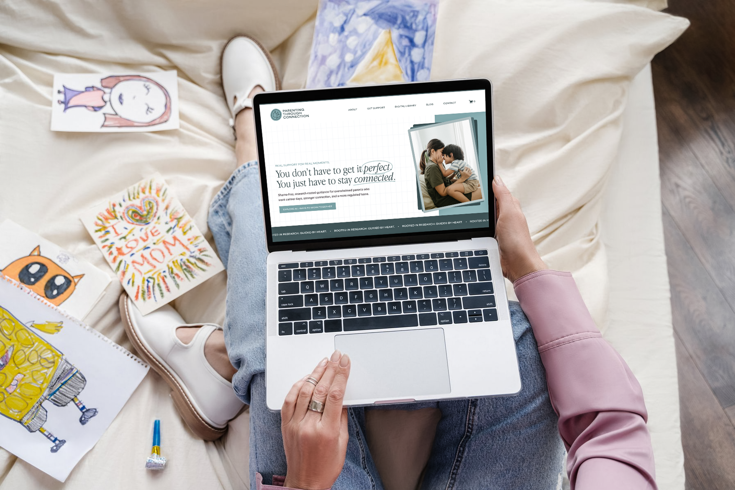 Person holding a laptop displaying a website called 'Parenting Through Connection' with a photo of a parent and child, surrounded by children's drawings and art supplies on a beige blanket.