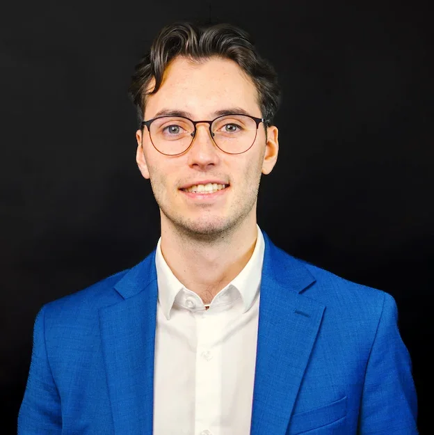 Un jeune homme portant un costume bleu, chemise blanche, lunettes, souriant, fond noir.