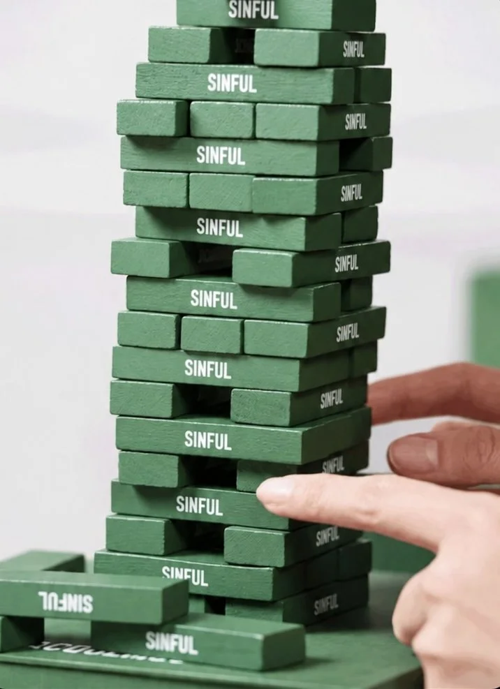 Green Jenga blocks with the word 'SINFUL' printed on each, stacked in a tower, with a hand about to remove one block.