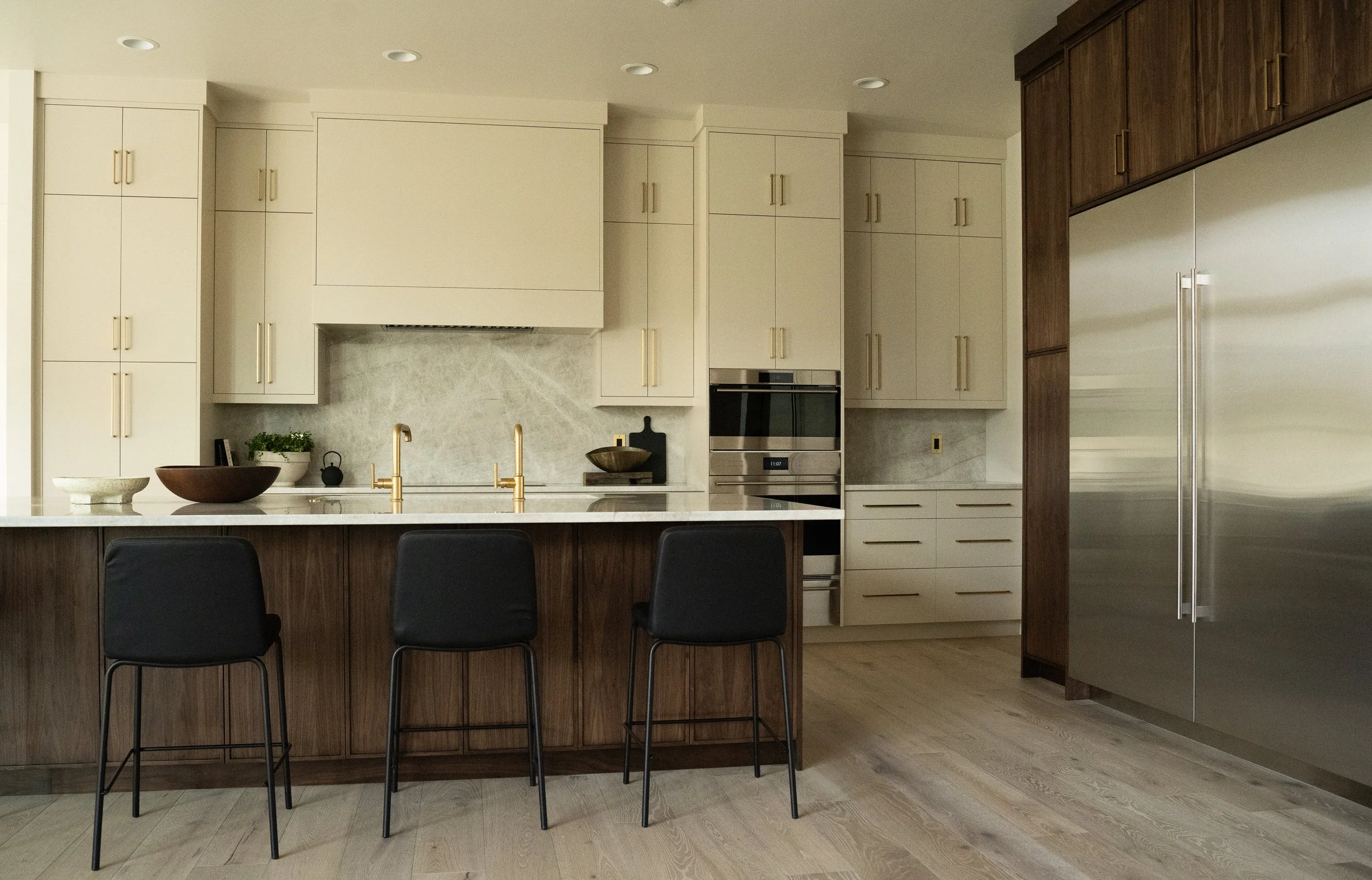 Modern kitchen with white cabinets, wooden accents, marble backsplash, stainless steel appliances, a kitchen island with black chairs, and decorative bowls and plants.