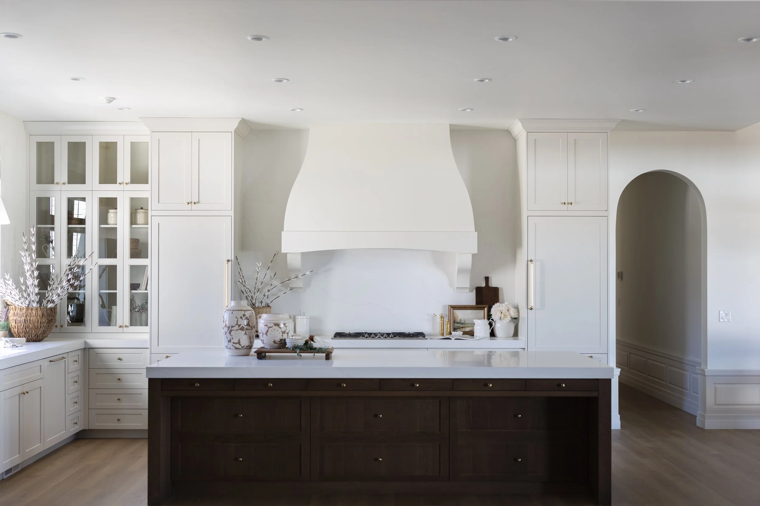 A modern kitchen with white cabinetry, a central dark wood island, decorated with vases and flowers, and a white range hood above a stove.