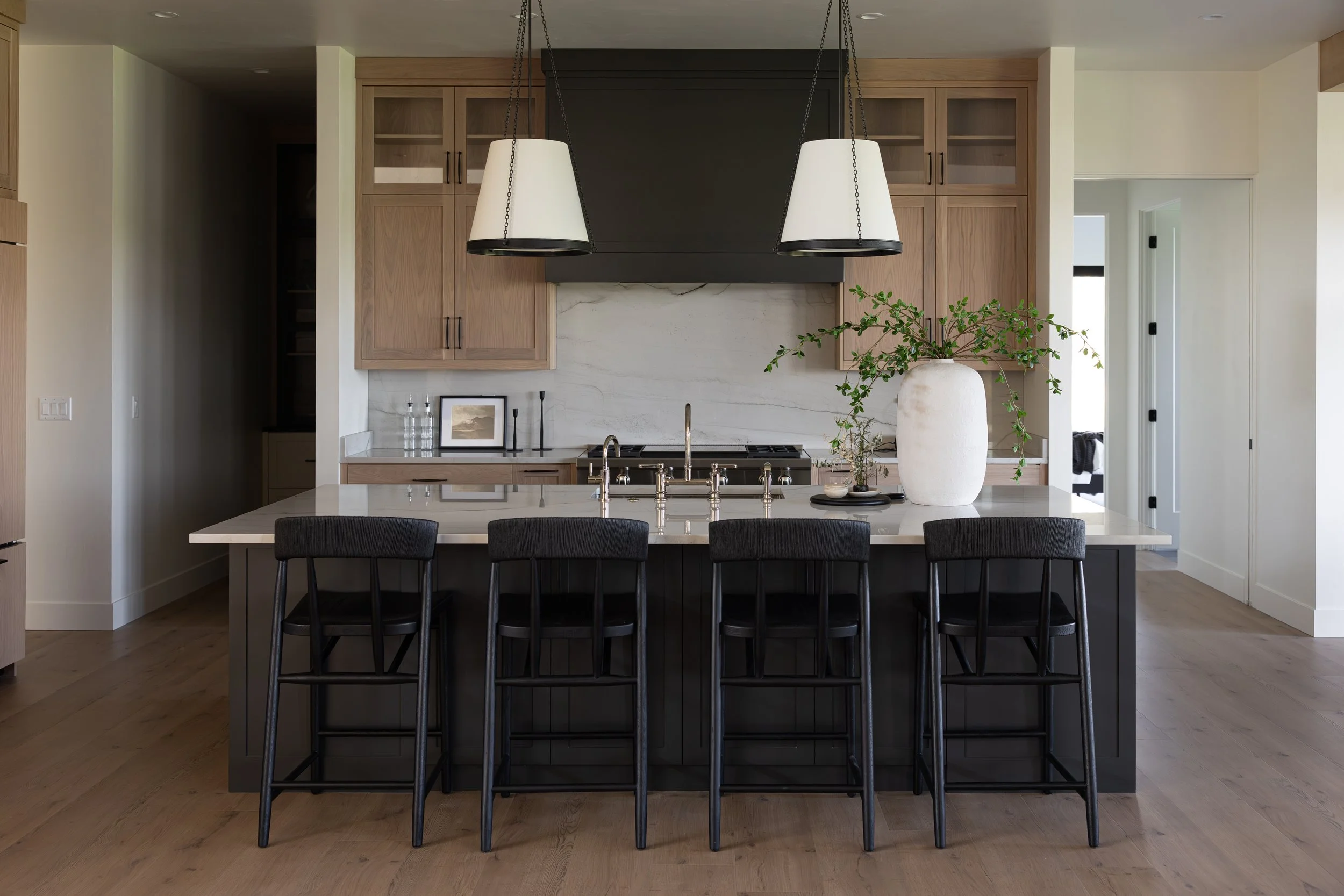 Modern kitchen with a large island featuring black chairs, wooden cabinets, a marble backsplash, pendant lighting, a large white vase with green foliage, and stainless steel sink.