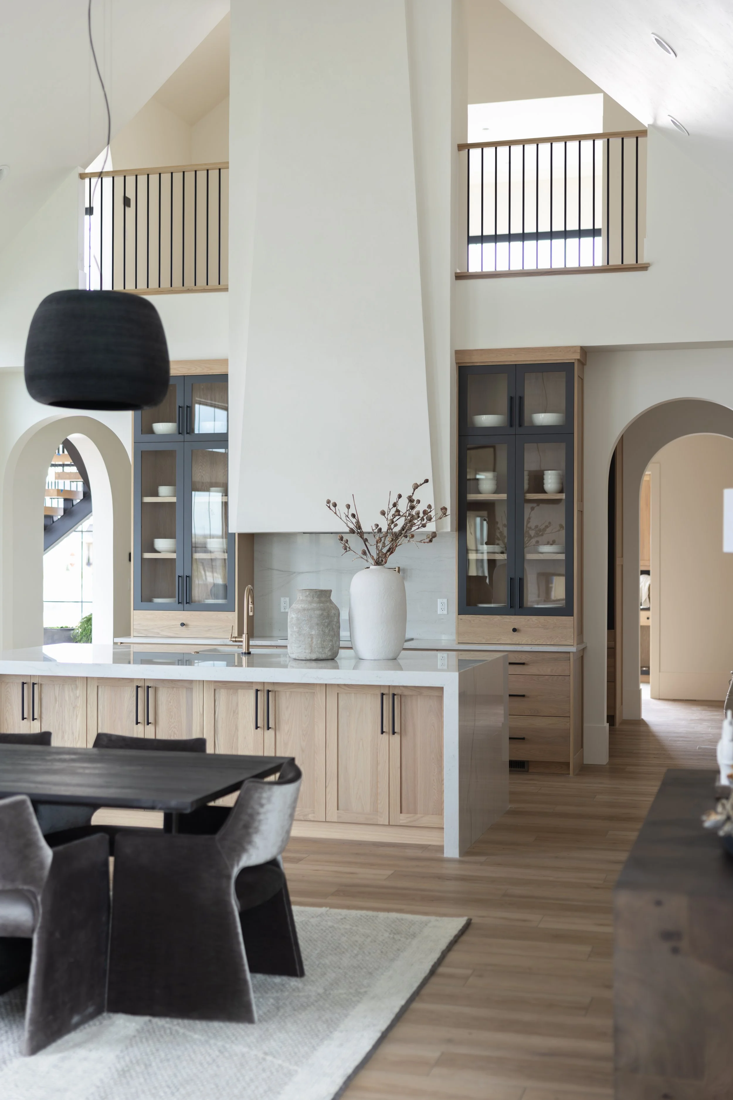 Open concept kitchen and dining area with light wood cabinets, a white kitchen island, black chairs, and decorative vases on the island. The space features high ceilings with skylights, a balcony with black railing, and soft neutral colors.