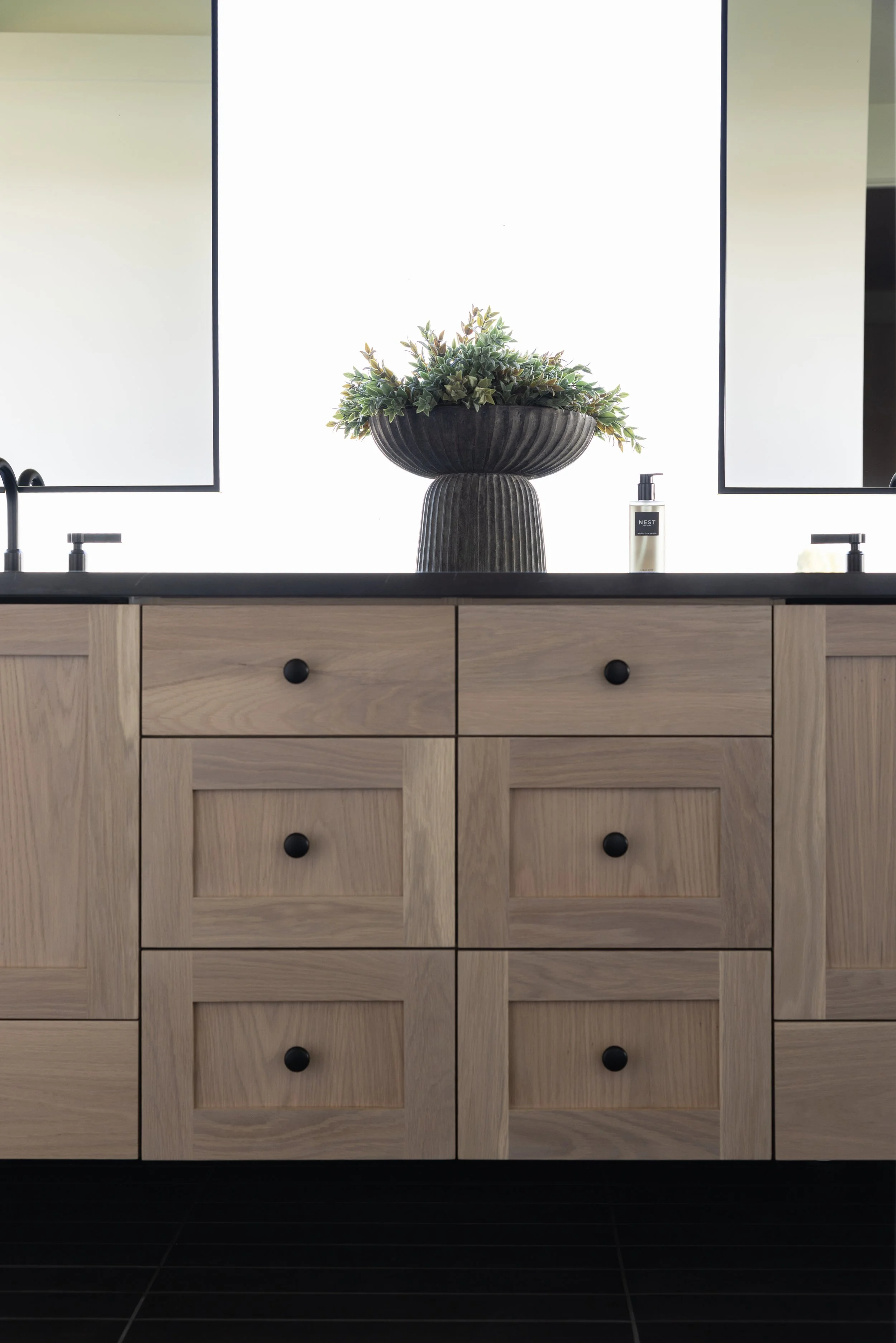 A bathroom vanity with a light wood finish, featuring two mirrors, a black faucet, a potted plant, and a bottle of NEST soap. The vanity has six drawers with black knobs, and the counter is black. The background includes a bright, backlit mirror or w