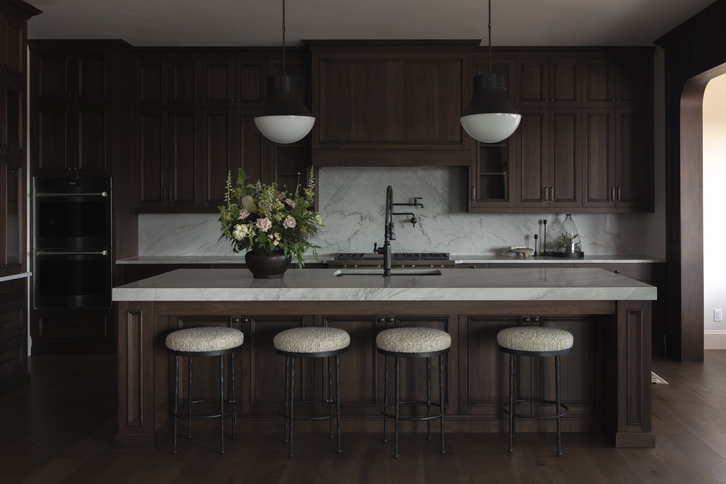 Modern kitchen with dark wood cabinets, white marble backsplash and island, black faucet, floral centerpiece on the island, and three barstools with woven beige seats.