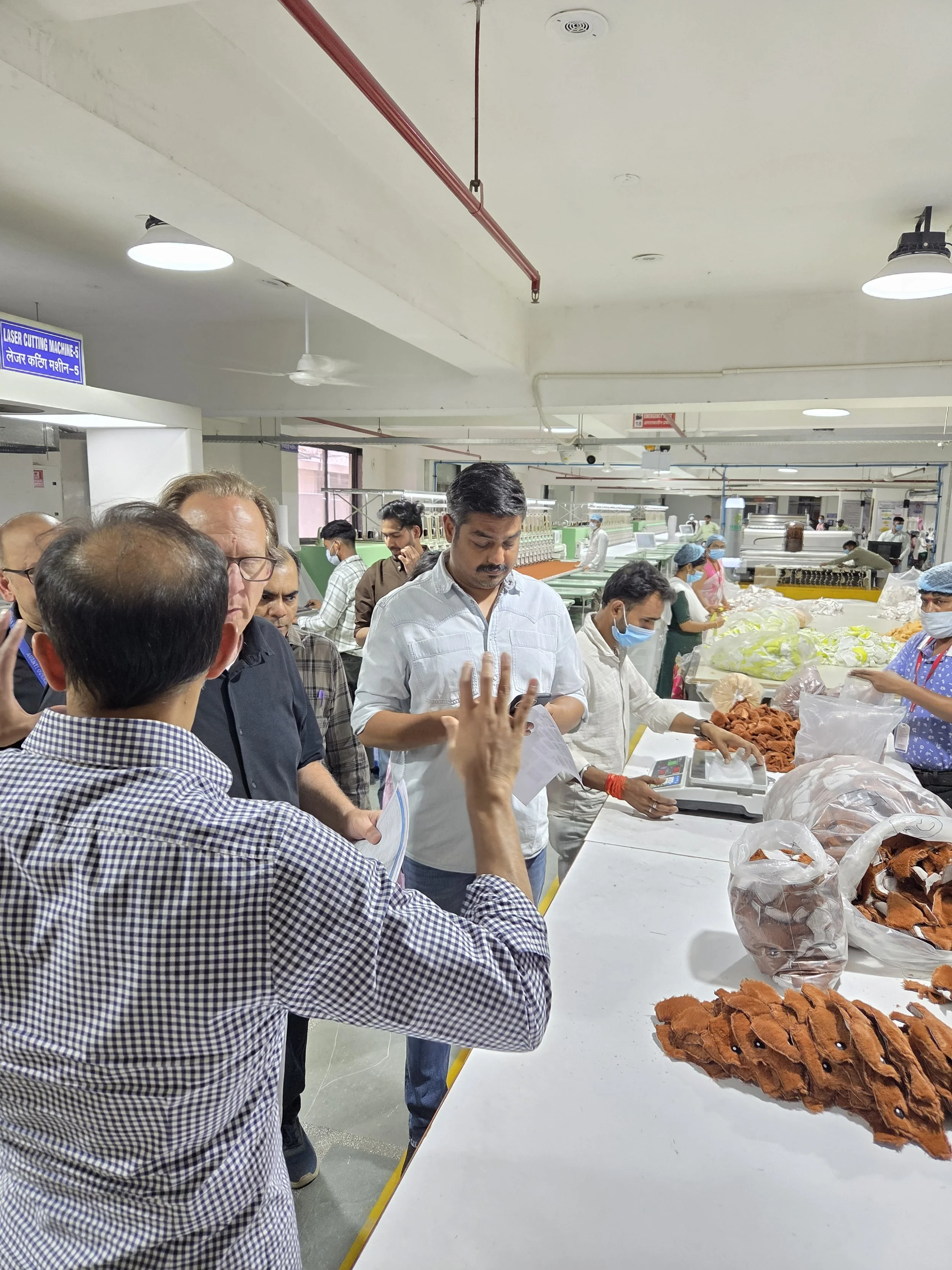 People in a factory or workshop inspecting and handling leather pieces, with some wearing masks, near workstations and equipment.