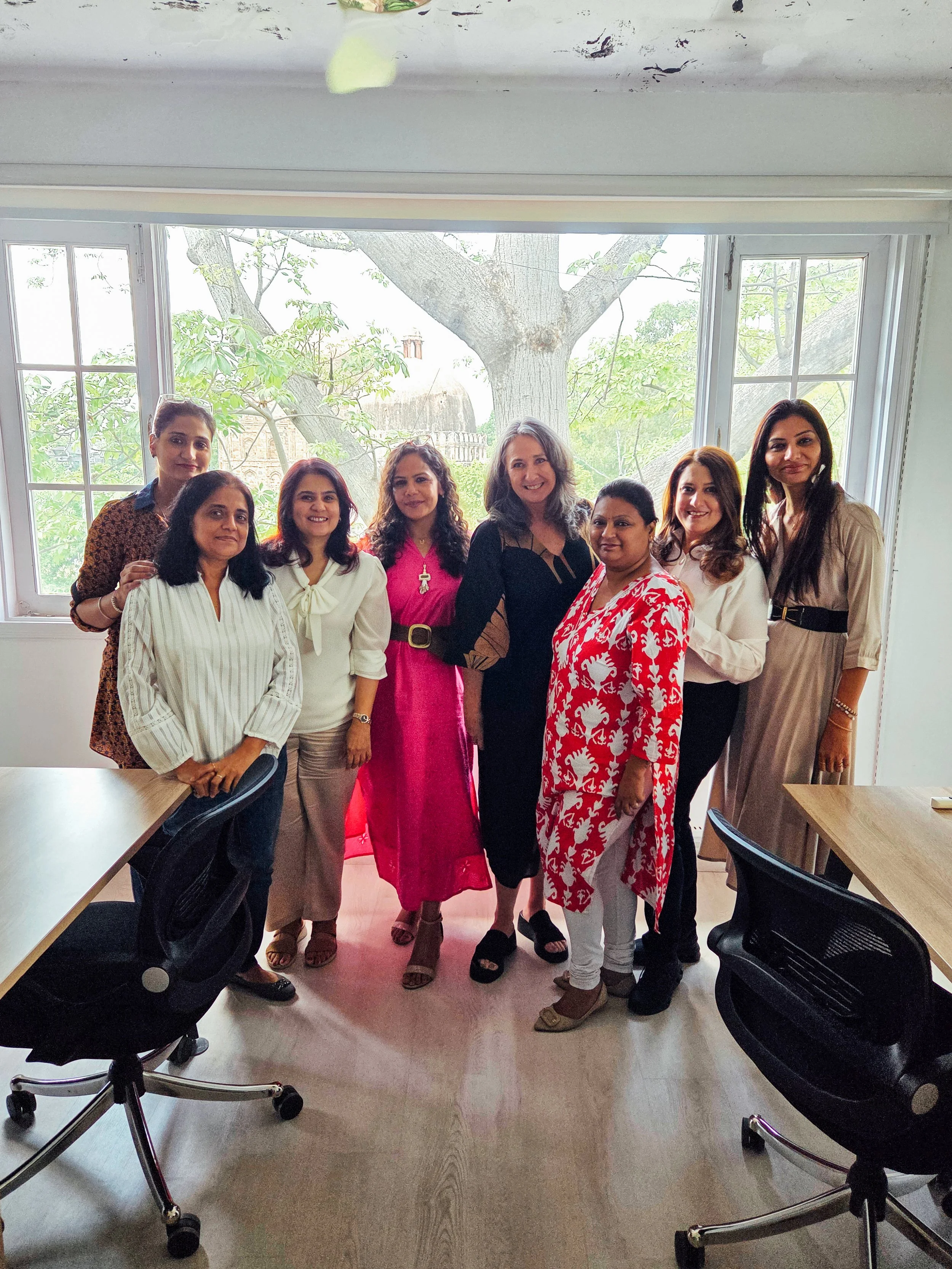 A group of nine women standing in a semi-circle in a room with large windows and a view of trees outside.