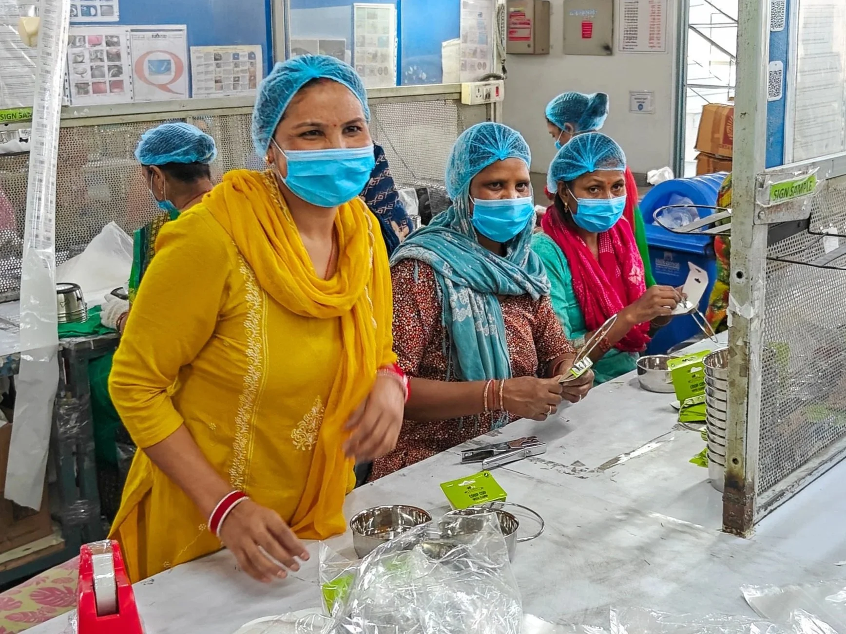 Group of women wearing blue hair nets and face masks working at a table in a manufacturing or packaging facility. They are handling small metal containers and miscellaneous items, with some products and supplies on the table in front of them.