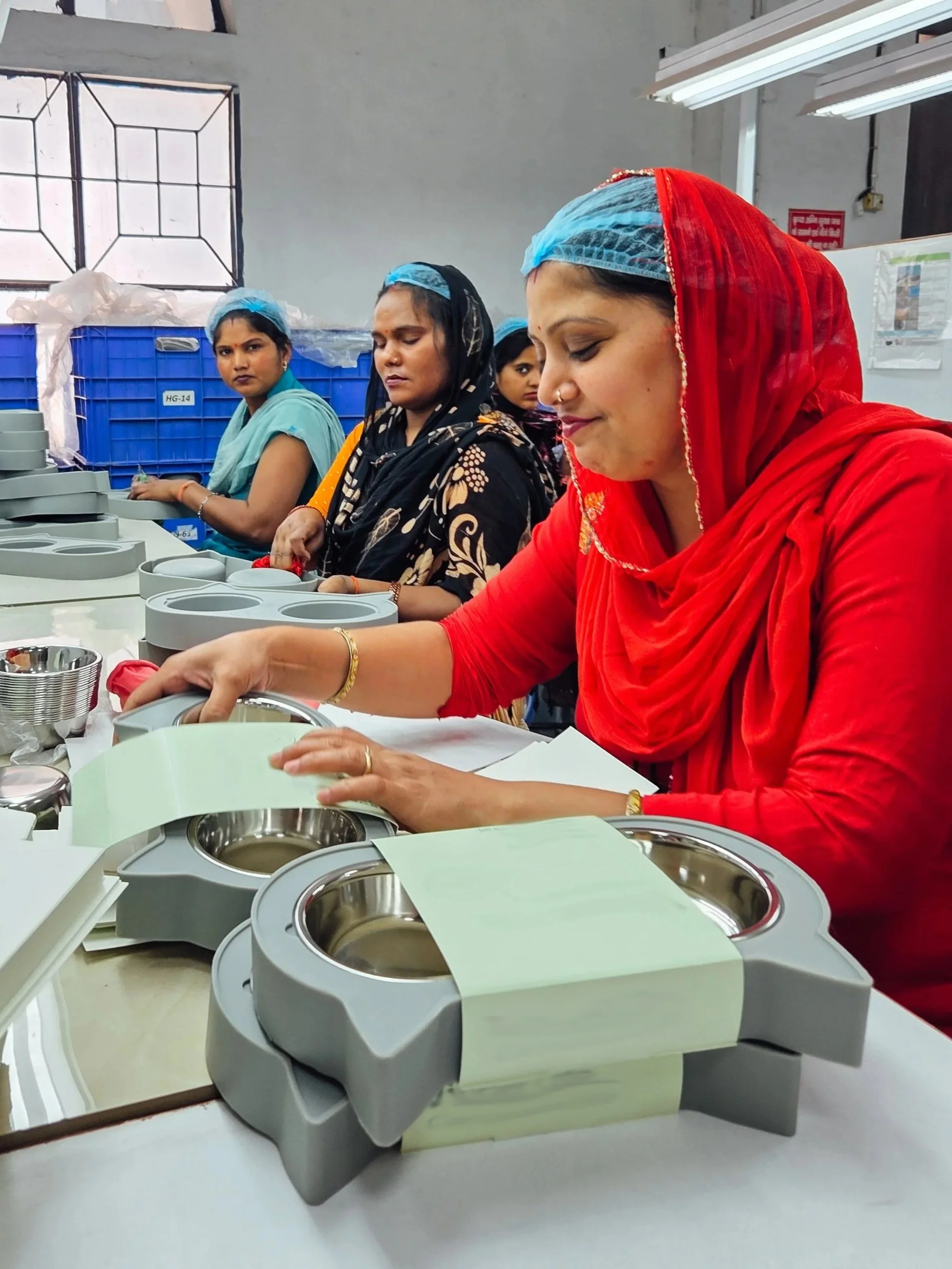 A group of women working with packaging of products in a room, with some women wearing traditional Indian attire and hair nets, likely involved in manufacturing process