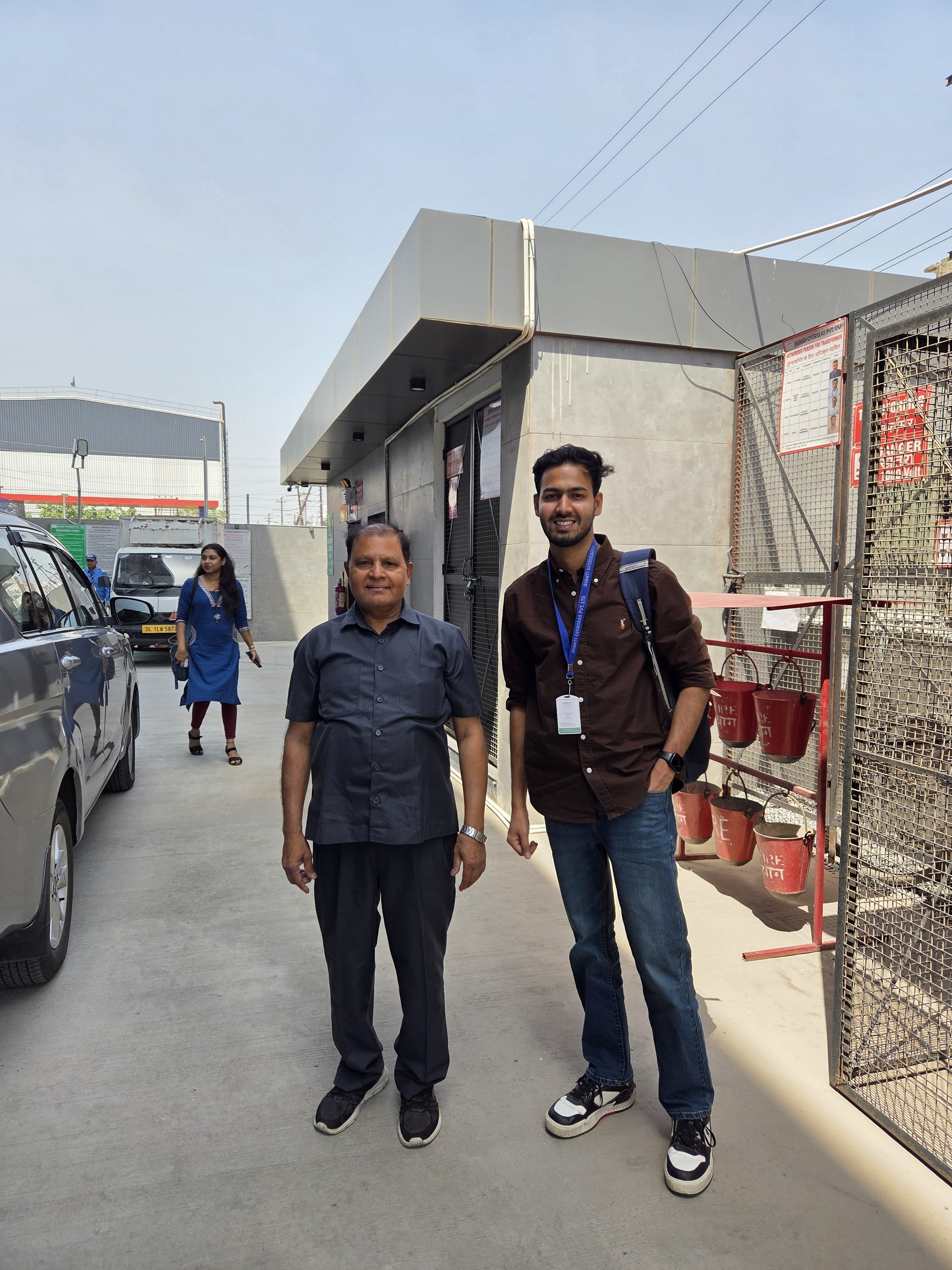 Two men standing outside a building, with one man in a black shirt and another in a brown jacket, surrounded by cars and people, with a clear blue sky overhead.