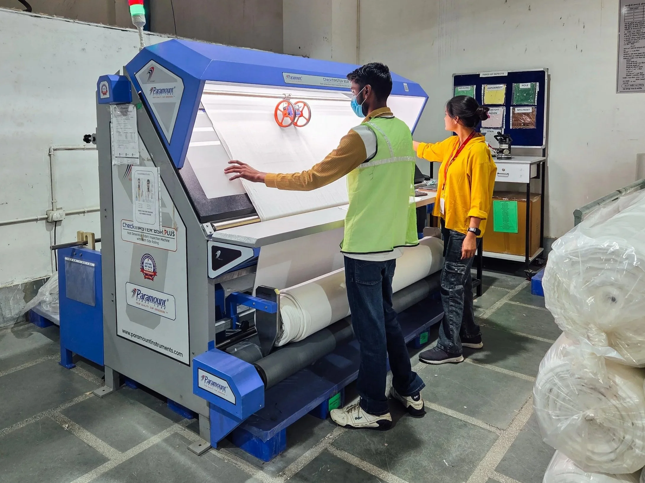 Two workers at a quality control station inspecting a rolled material on a conveyor in an industrial setting.