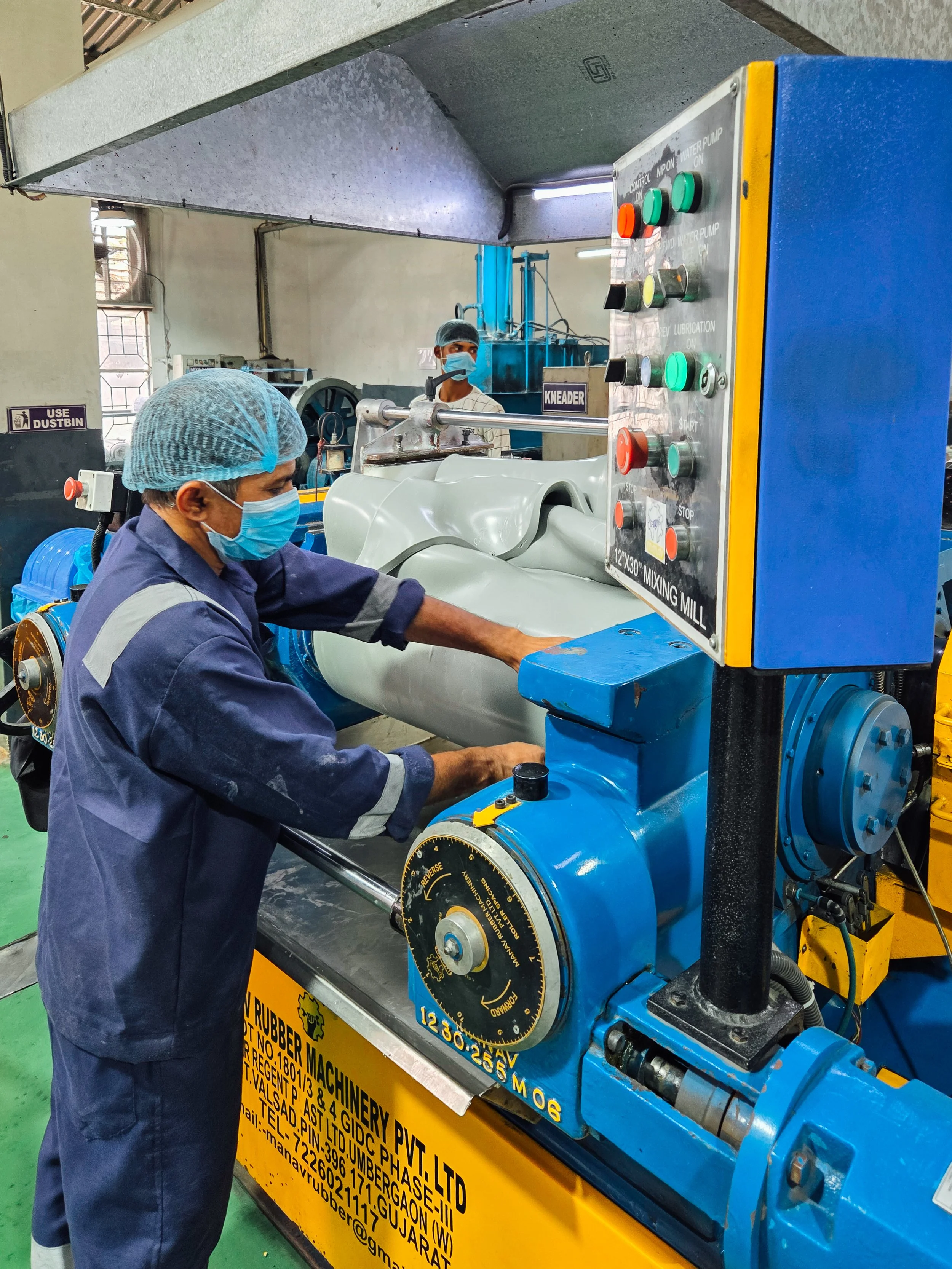 Two workers in safety masks and hairnets operating a blue industrial mixing mill machine inside a factory. One worker is adjusting the machine while the other observes in the background.
