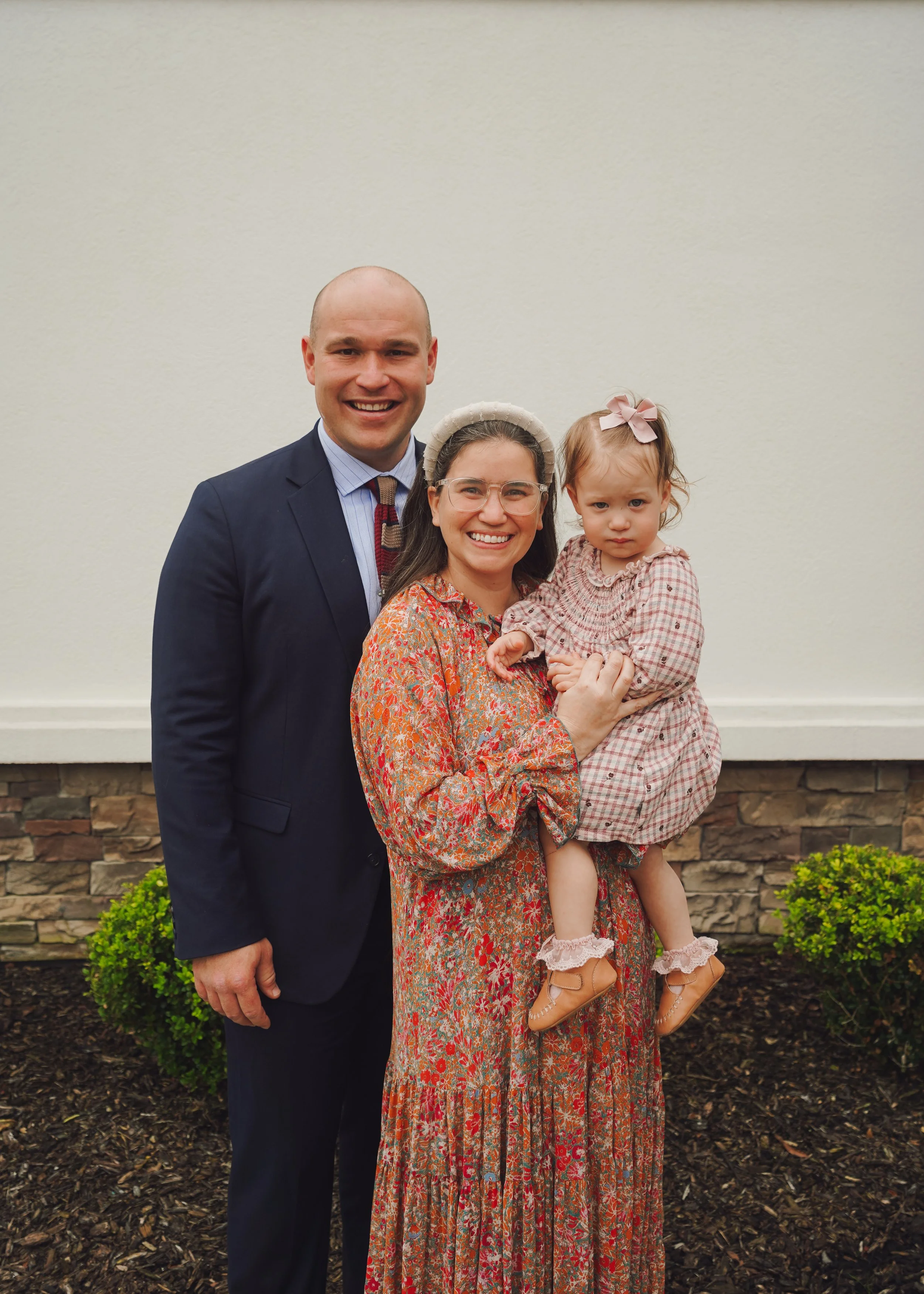 A family of three, including a man, woman, and young girl, standing outside against a beige wall and dark mulched soil with green bushes, smiling and posing for a photo.