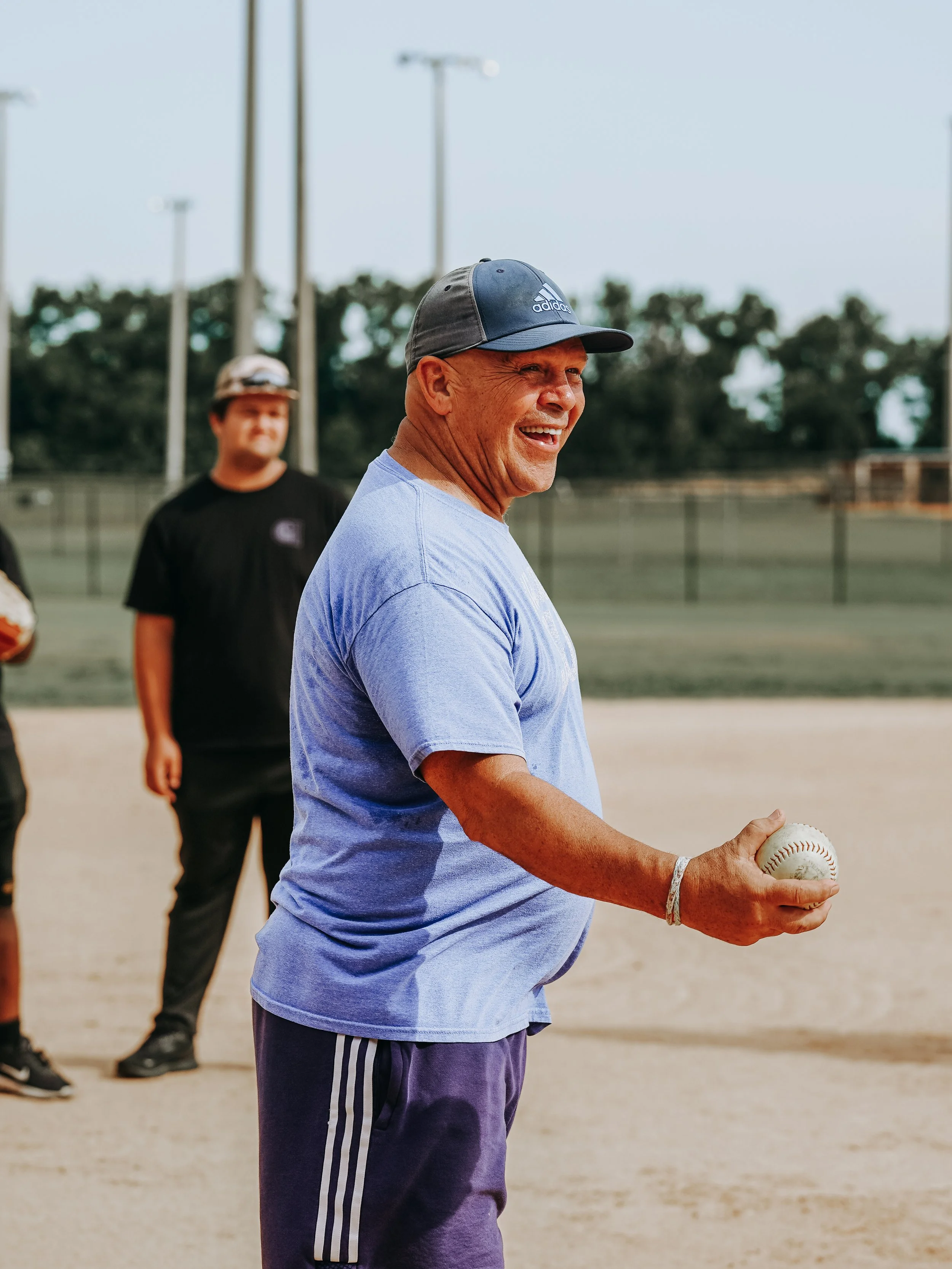 A smiling elderly man wearing a blue cap and a blue t-shirt holding a baseball at a baseball field.
