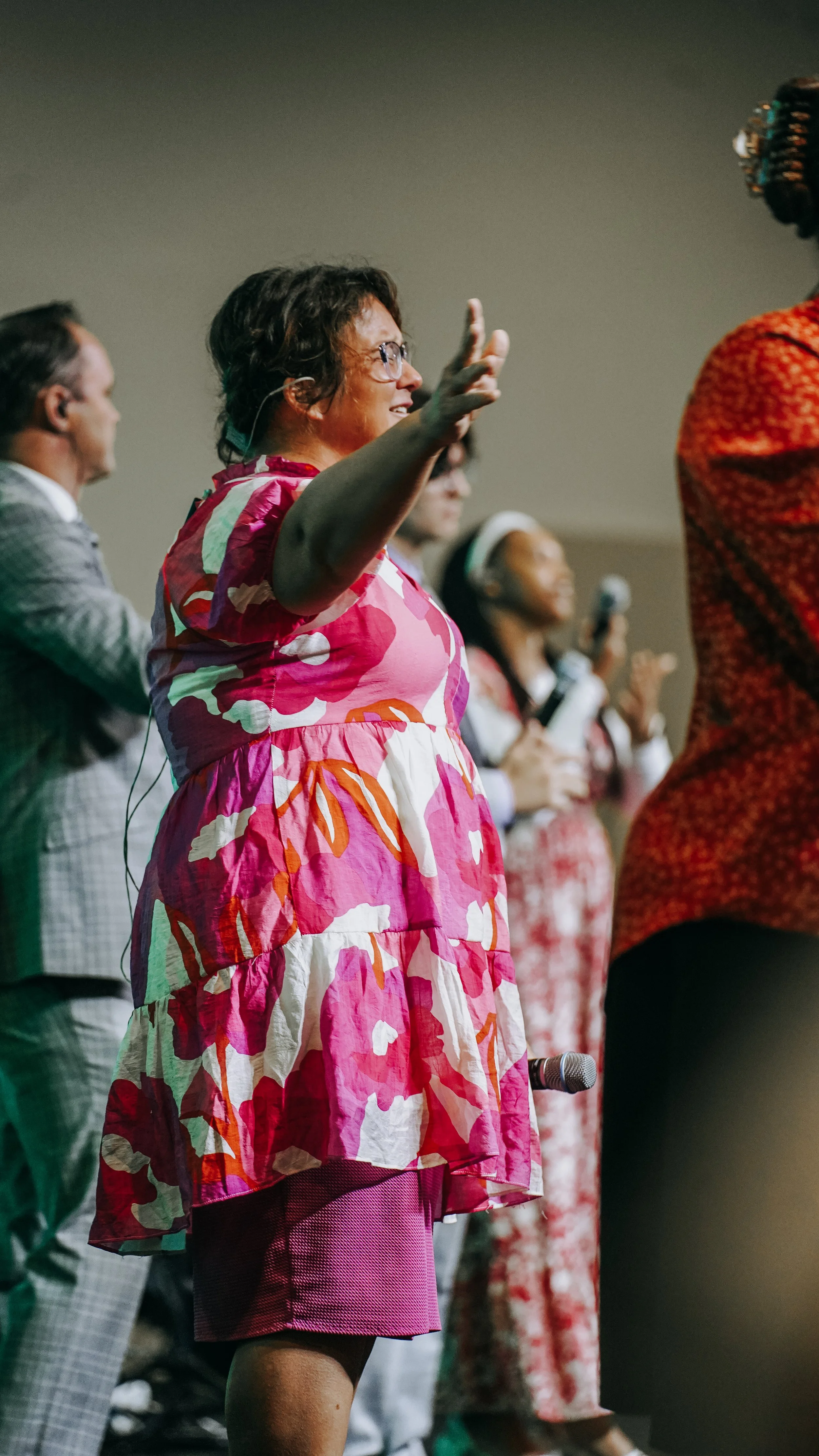 A group of diverse people on stage during a presentation or performance, with one woman in a colorful pink and red dress in the foreground.