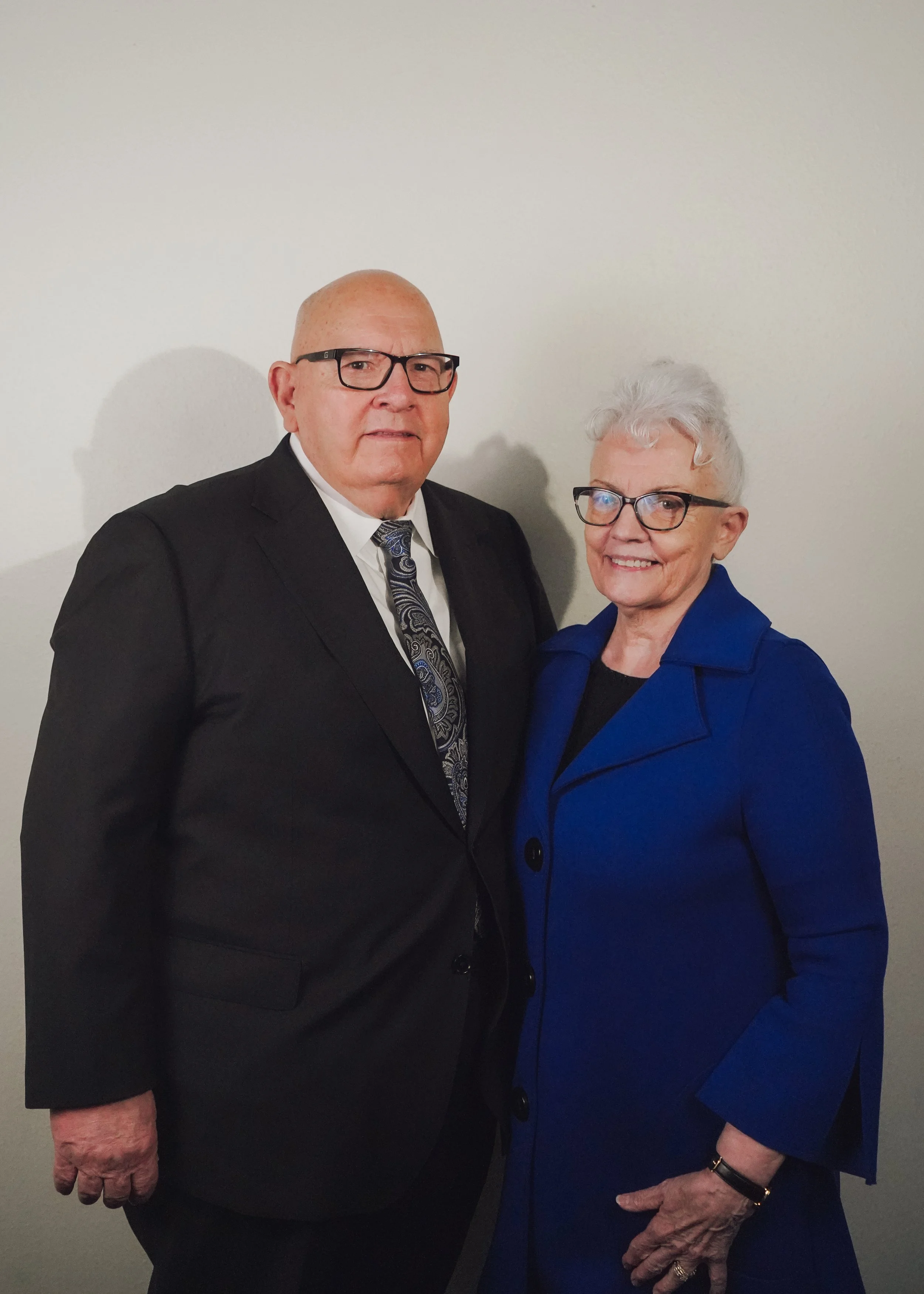 A man in a dark suit, patterned tie, and glasses standing next to a woman in a blue blazer, glasses, and white hair, both smiling, against a plain light-colored wall.