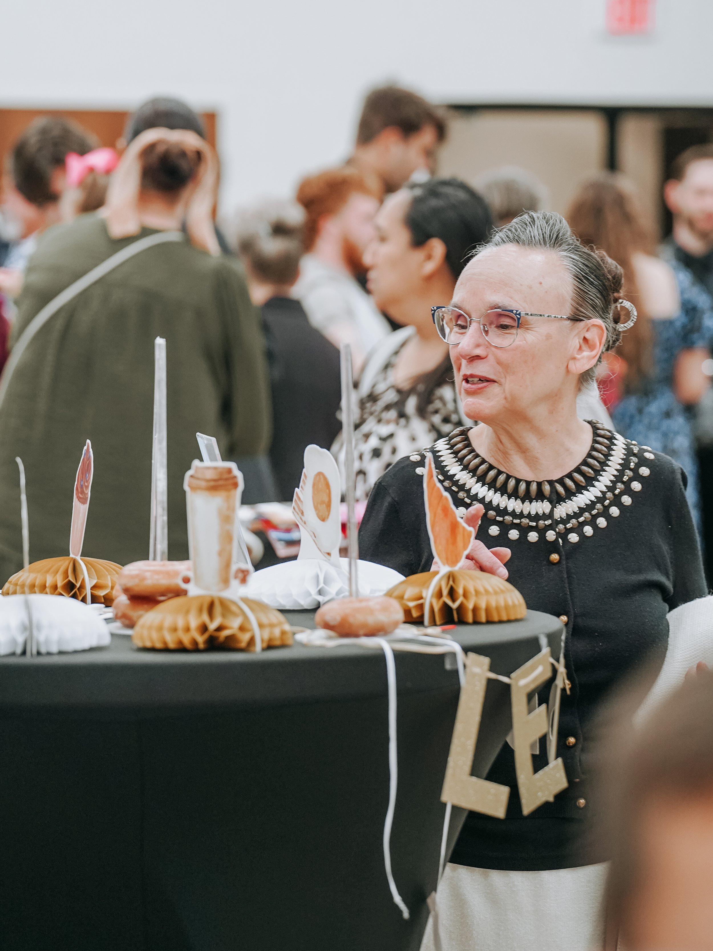 A group of people at a social gathering or celebration, with a woman in the foreground wearing glasses, a black dress with pearl embellishments, and a necklace, sitting at a table with decorative items and small flags. Other people are visible in the background, engaging and mingling.