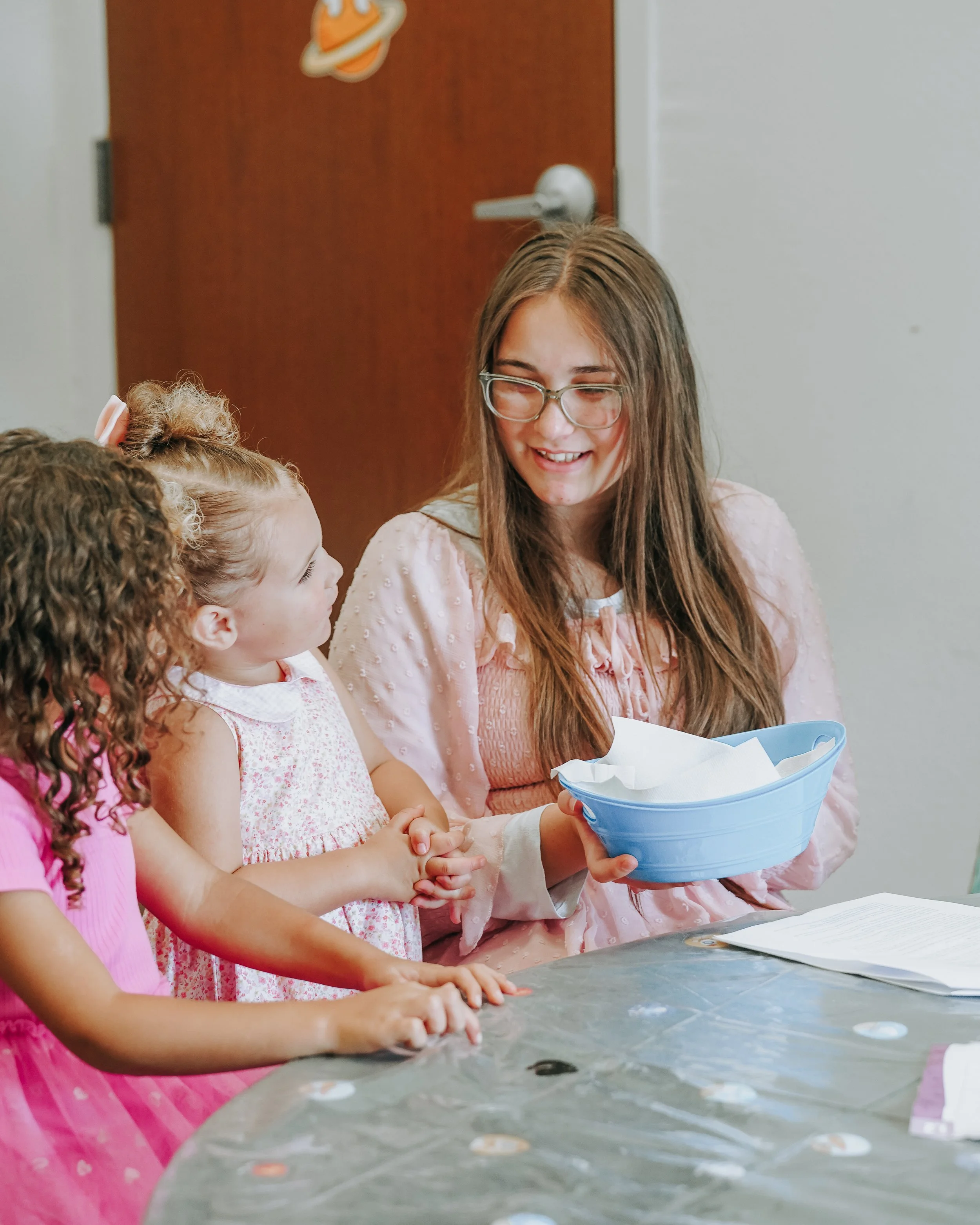 A young woman with long brown hair, glasses, and a light pink dress holding a blue bowl with tissues, engaging with children at a table in a classroom or activity setting.