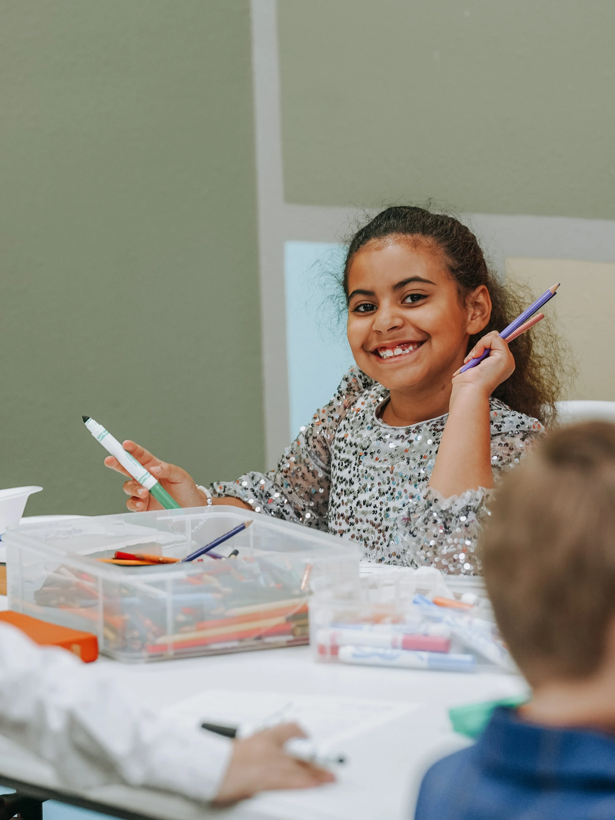 A young girl smiling and holding a purple pen during a classroom activity, with other children and art supplies on the table.