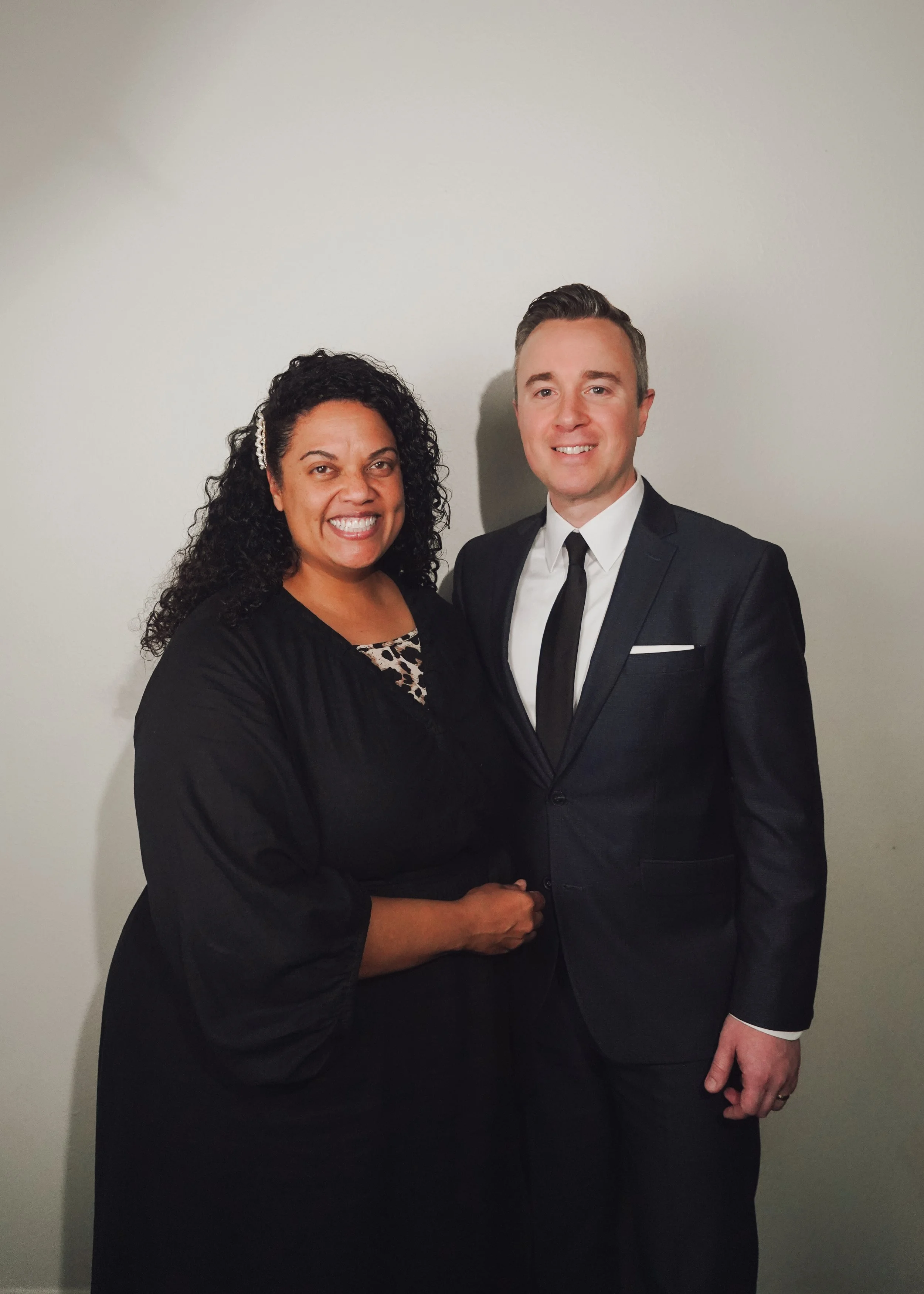 A woman and a man are standing close together, smiling, and shaking hands against a plain light-colored wall. The woman has curly black hair and is wearing a black dress, while the man has short, neatly styled hair and is dressed in a black suit with a white shirt and black tie.