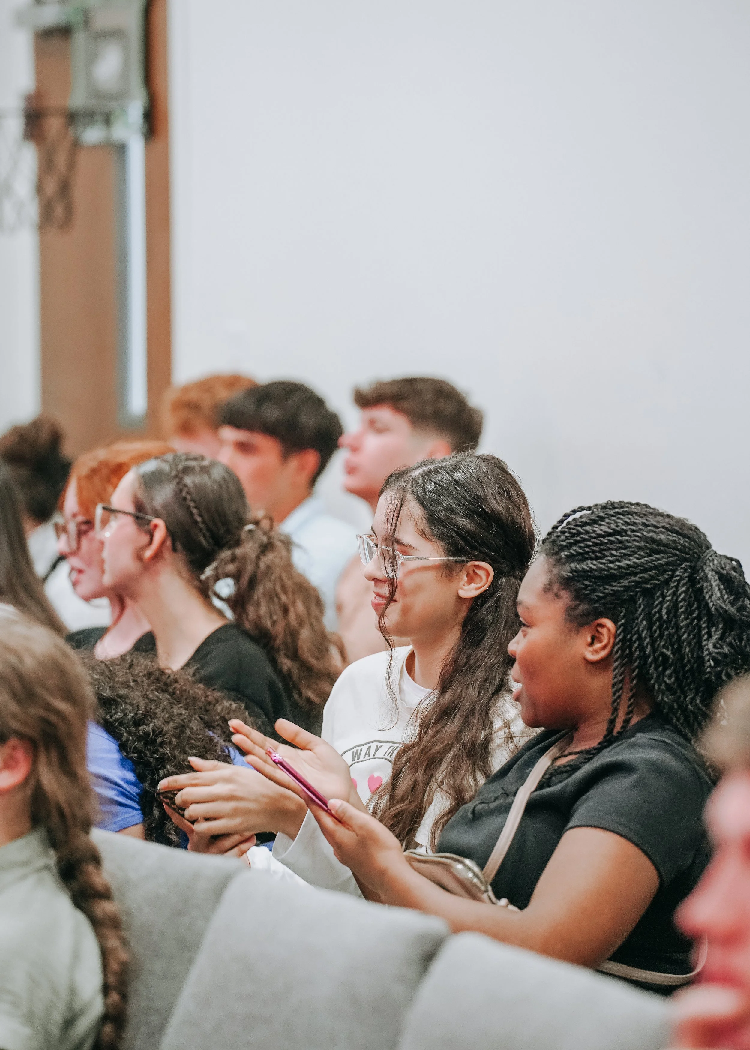 A diverse group of people sitting in chairs at a conference or event, some taking notes, with a plain white wall in the background.
