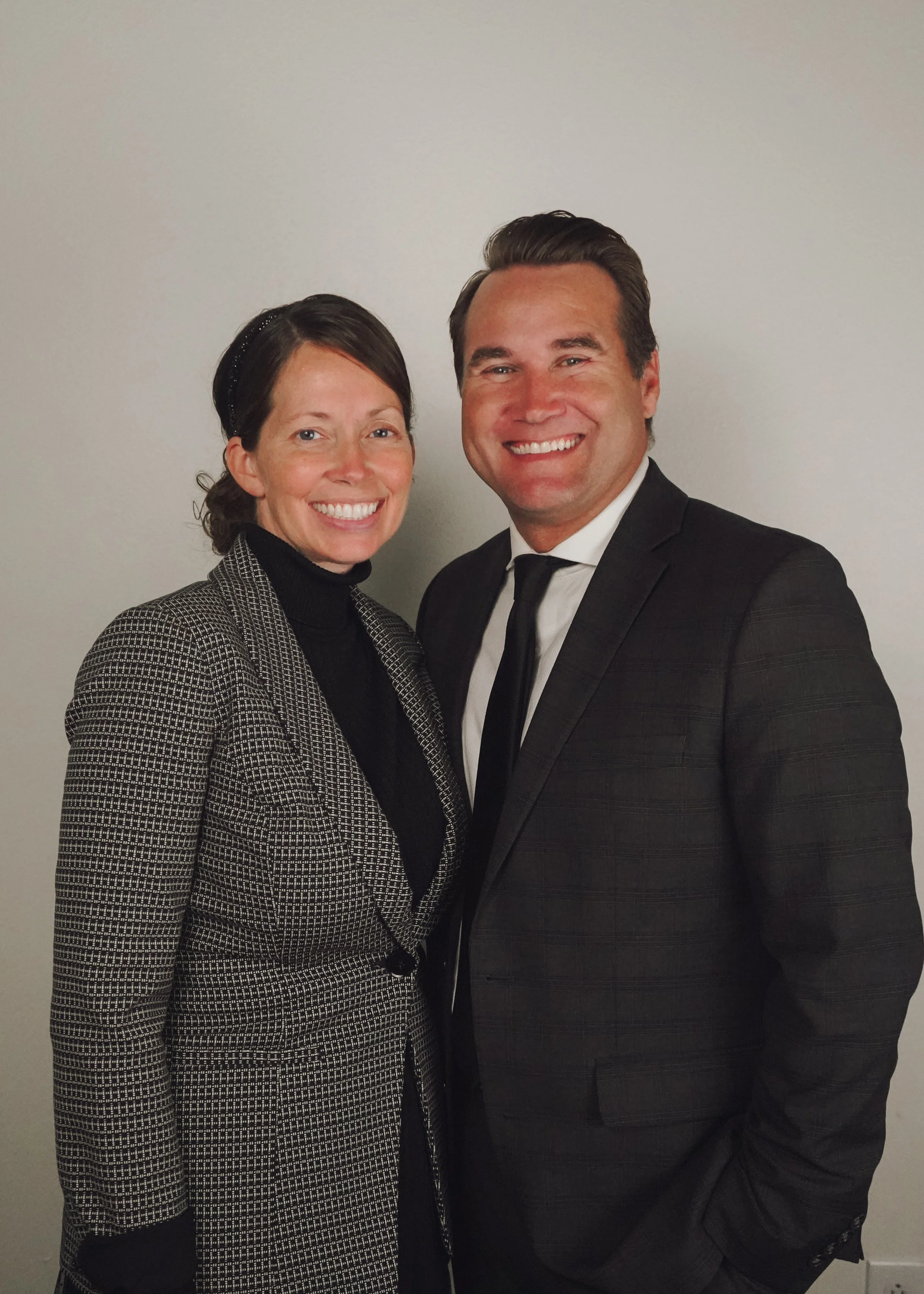 A smiling woman with dark hair wearing a black turtleneck and black-and-white patterned blazer standing next to a smiling man with dark hair combed back wearing a black suit, white shirt, and black tie, against a plain light background.