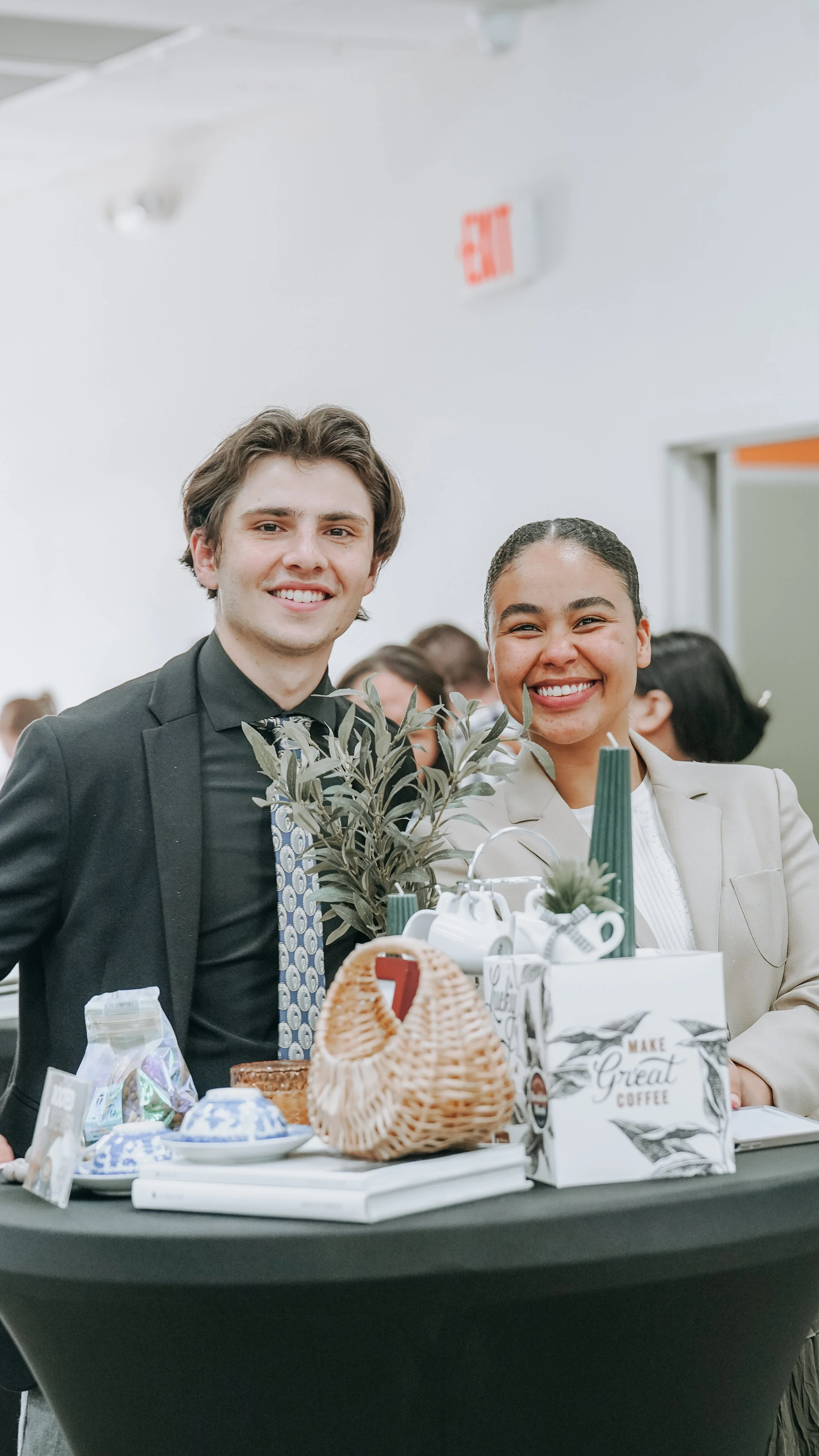 Two people, a man and a woman, smiling at an indoor event, standing behind a table with decorative items, plants, and small gift bags.