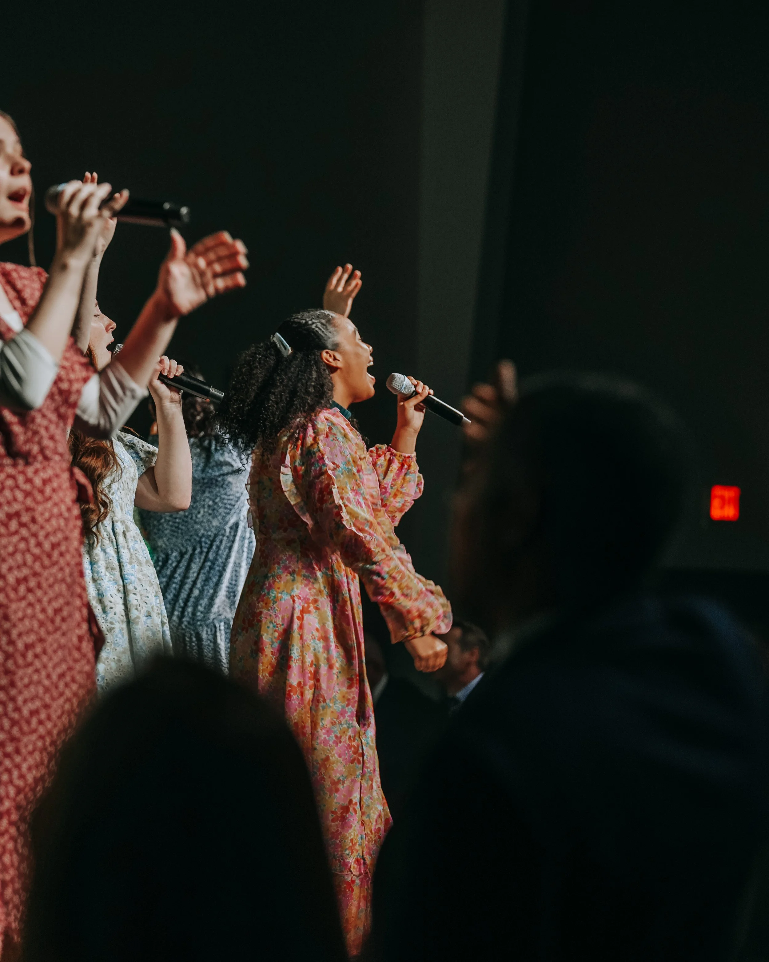 Women singing into microphones during a performance or event, with several women visible, some raising their hands, on a dimly lit stage.