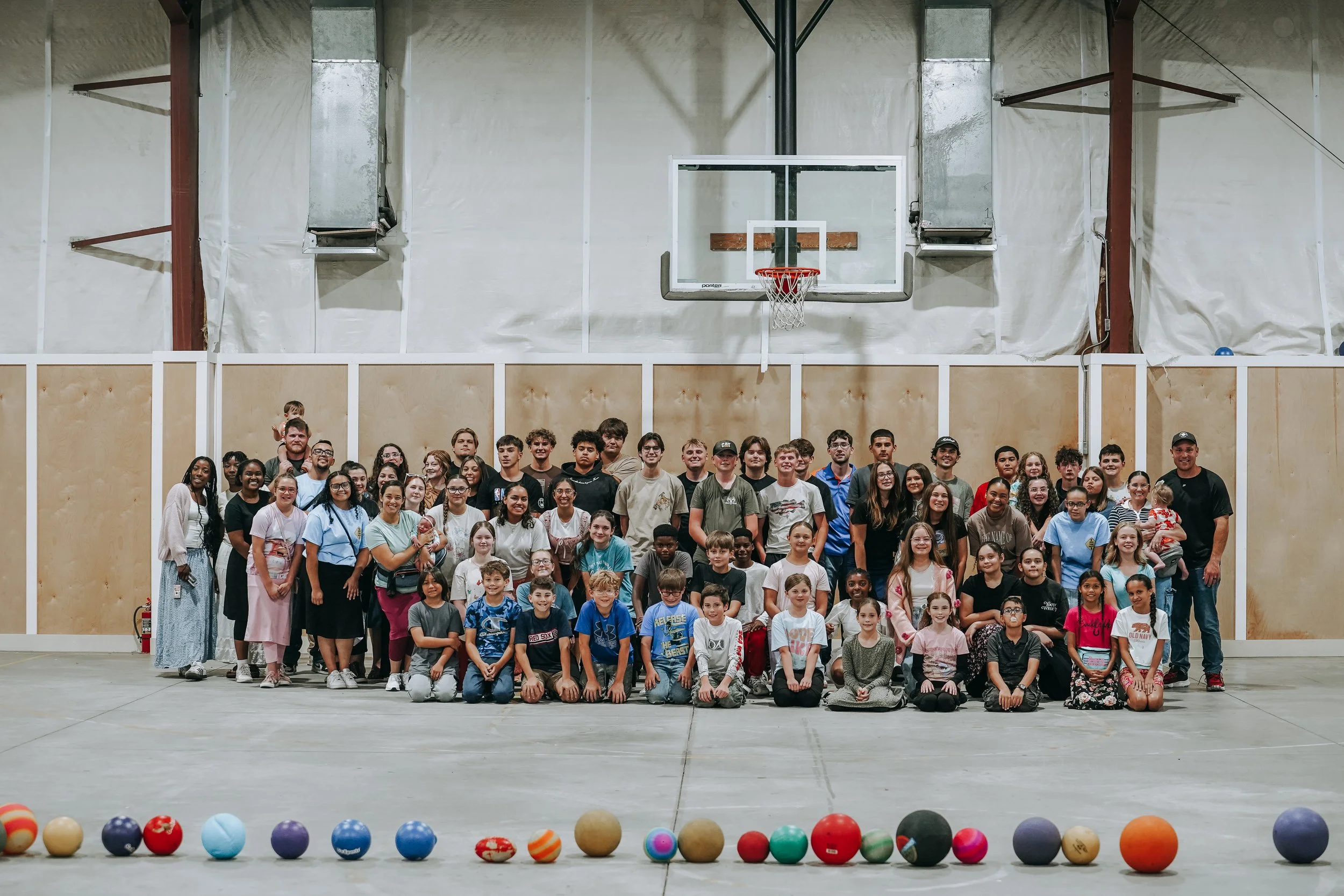 A large group of children and adults are gathered in a gymnasium, posing for a group photo in front of a basketball hoop. The foreground features a line of colorful balls on the floor.