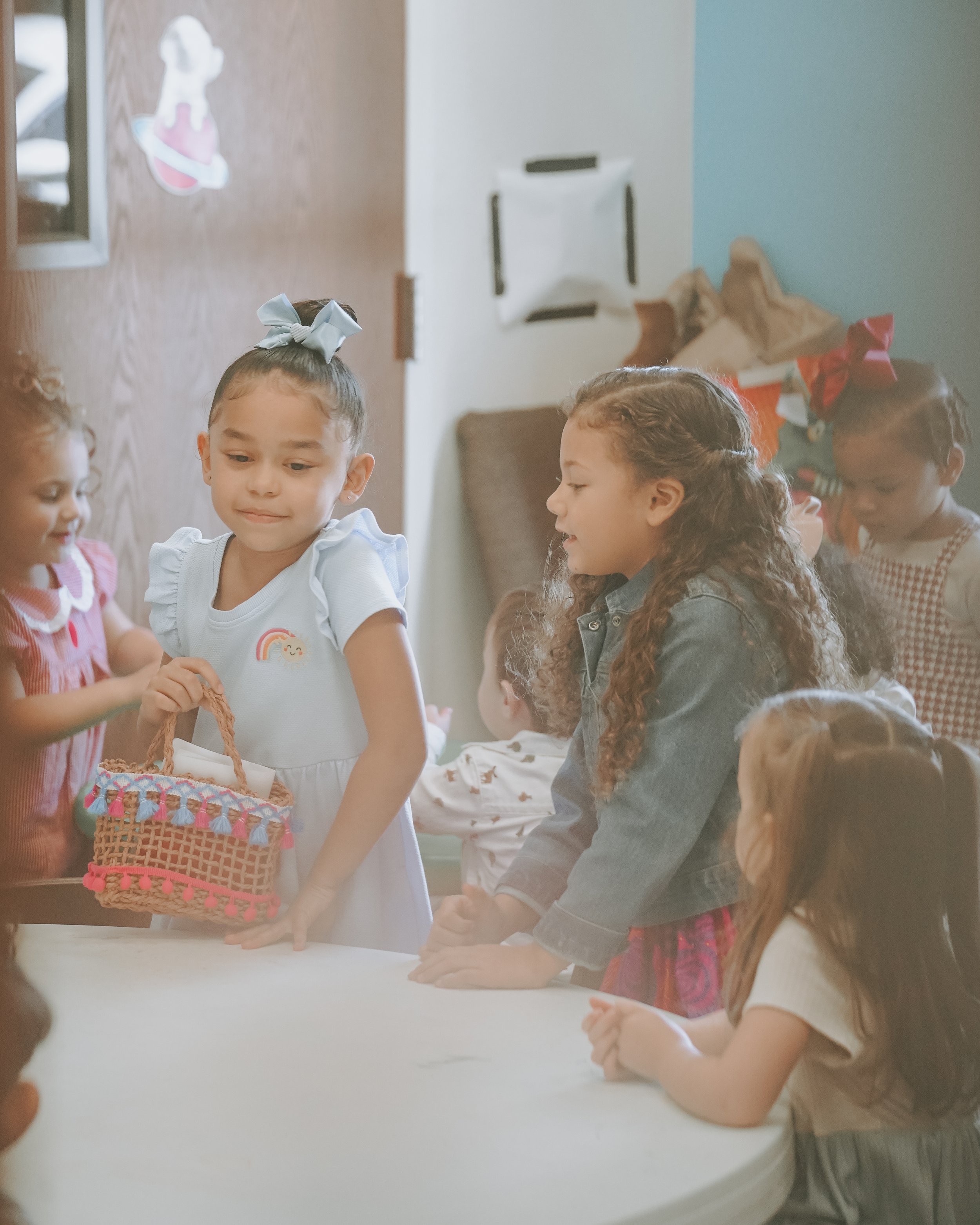 Group of young girls gathered around a table, some looking down, with a girl in the center holding a woven basket with tassels, inside a room decorated with a sticker of a dog on the wall.