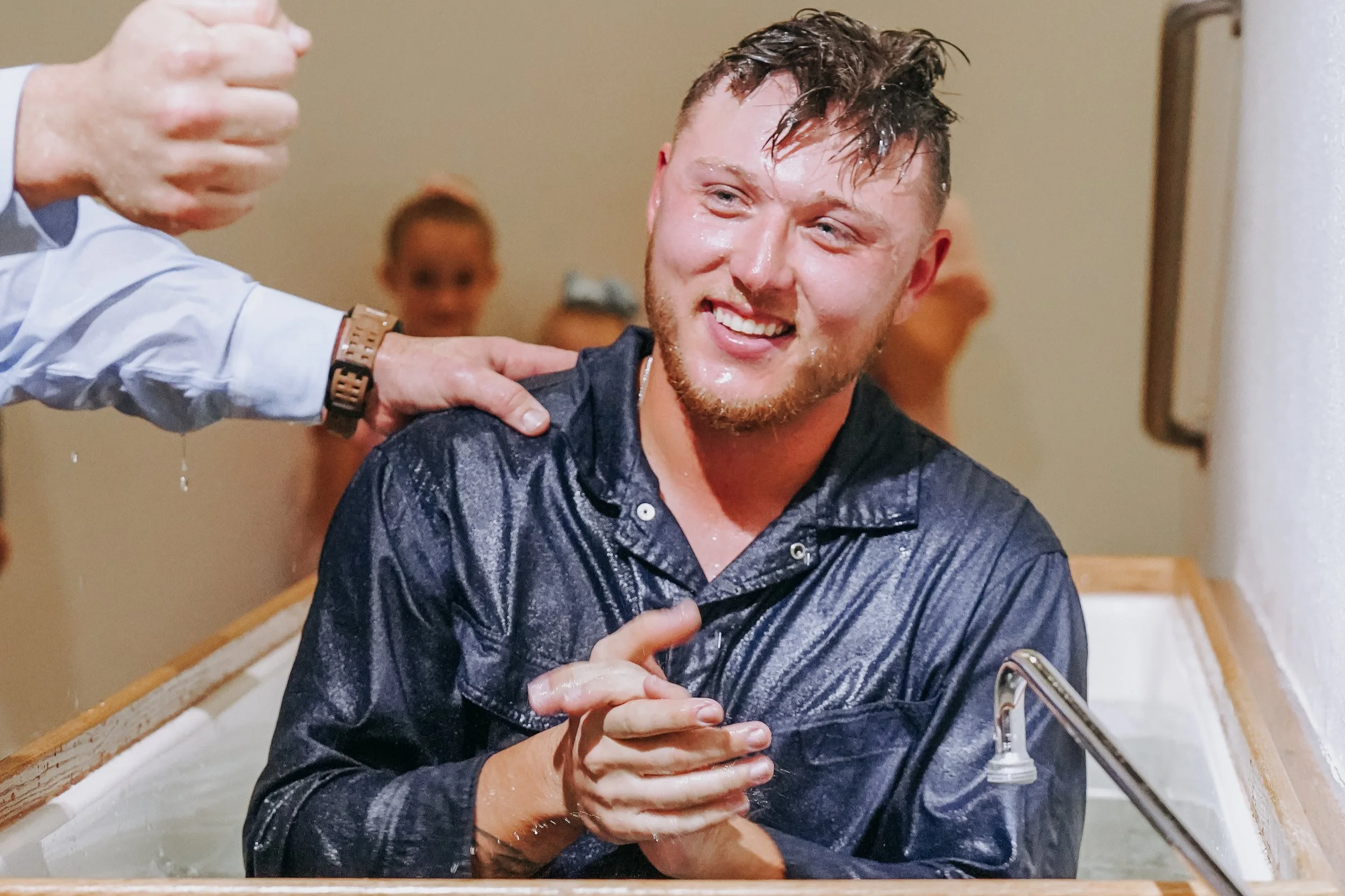 A young man with wet hair and a smiling expression is sitting in a sink as water is poured on his head by a person wearing a watch, in a bathroom. Two other children are blurred in the background.