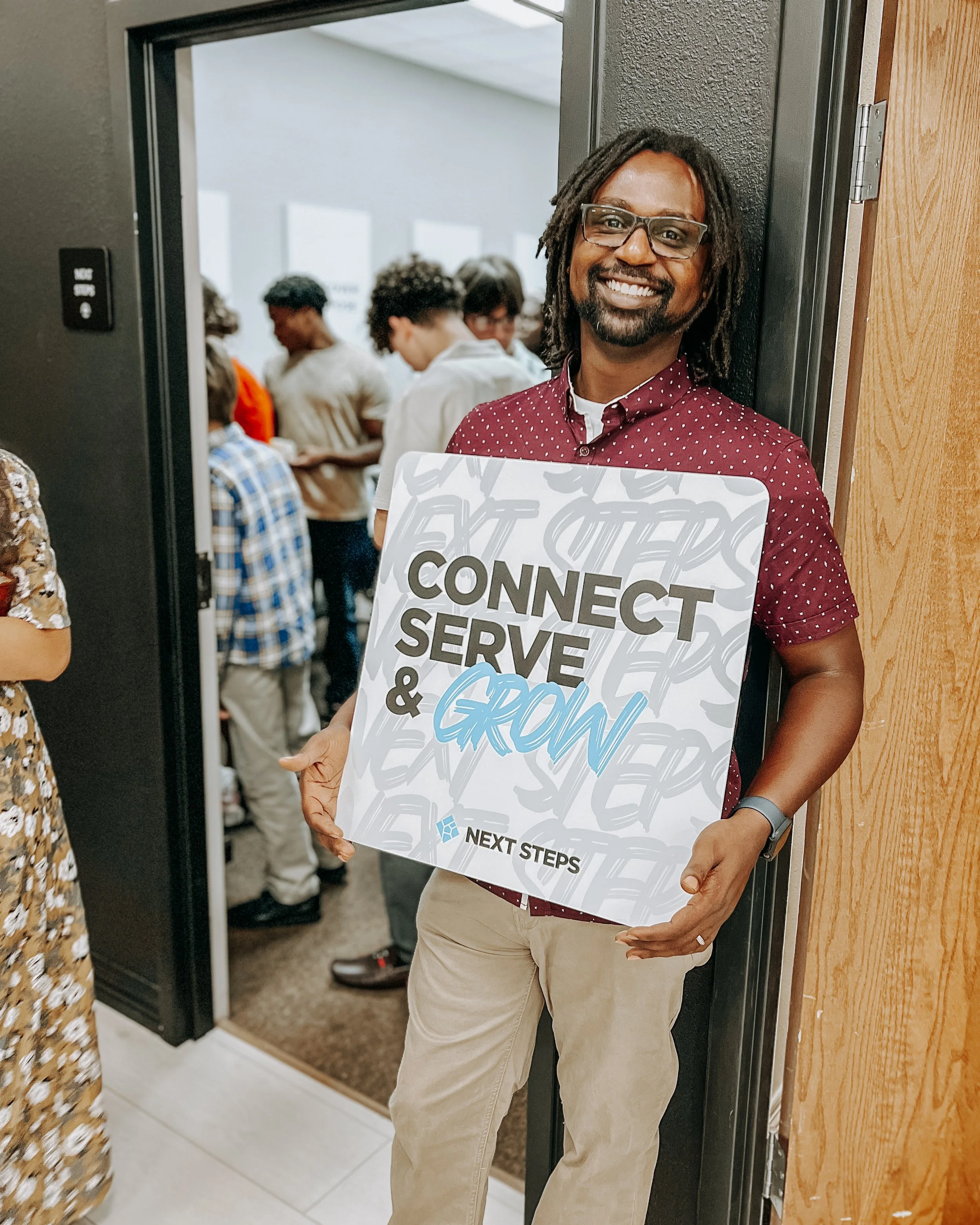 Smiling man with glasses holding a sign that reads "Connect, Serve & Grow" at an indoor event, with people in the background.