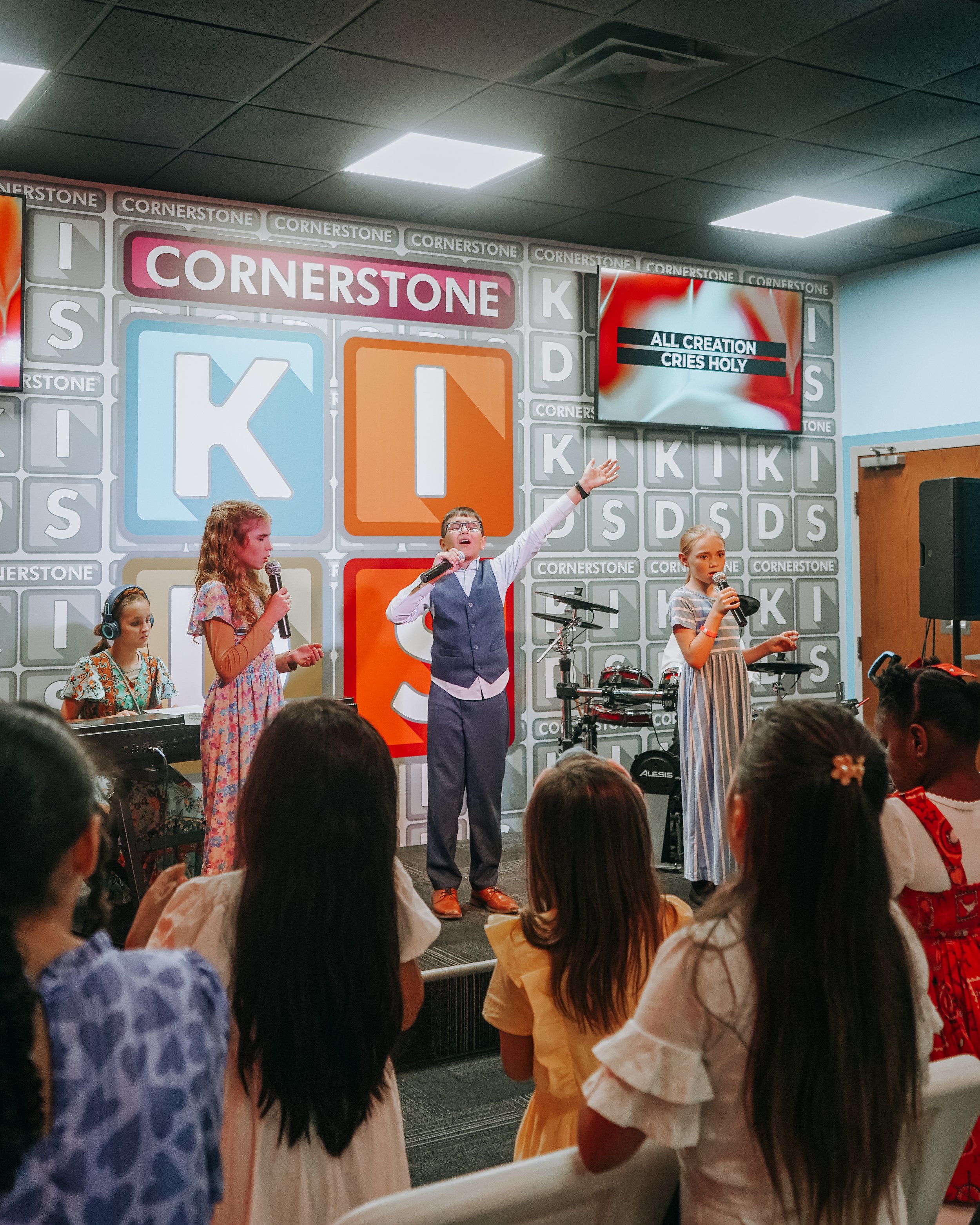 A group of children performing on stage at a church or school, singing into microphones, with a music keyboard and drums in the background, and an audience watching.
